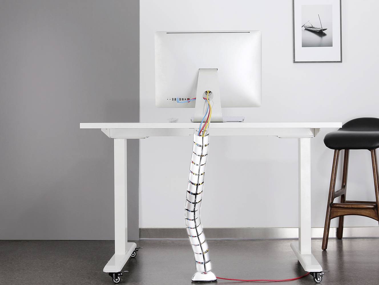 Ein Schreibtisch mit einem Computer, Kabelmanagement unterhalb des Tisches. Rechts ein Barhocker; an der Wand hängt ein gerahmtes Foto.