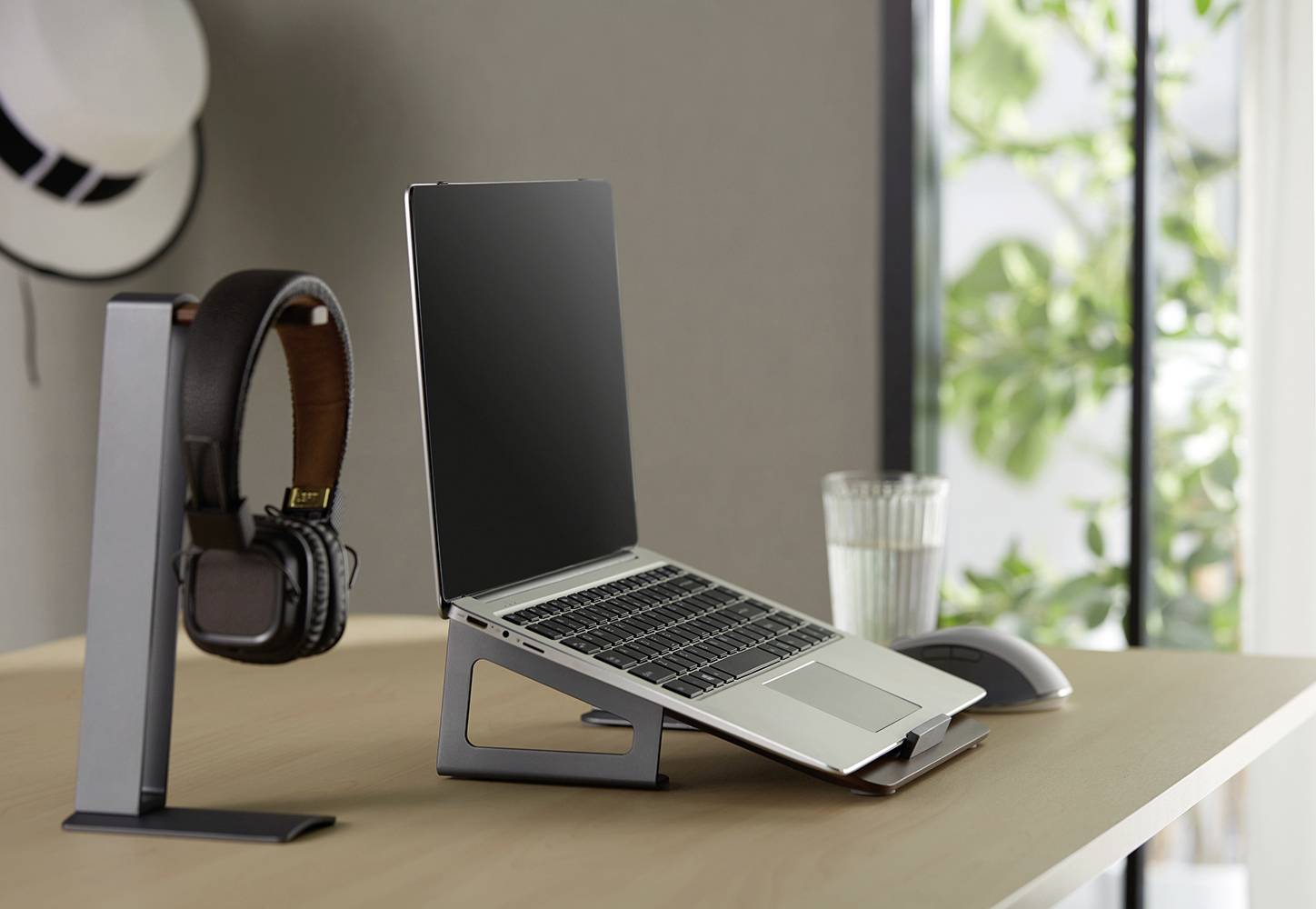 Ein Laptop auf einem Tisch steht auf einem Ständer. Daneben befinden sich Kopfhörer an einem Halter, ein Glas Wasser und eine Maus.