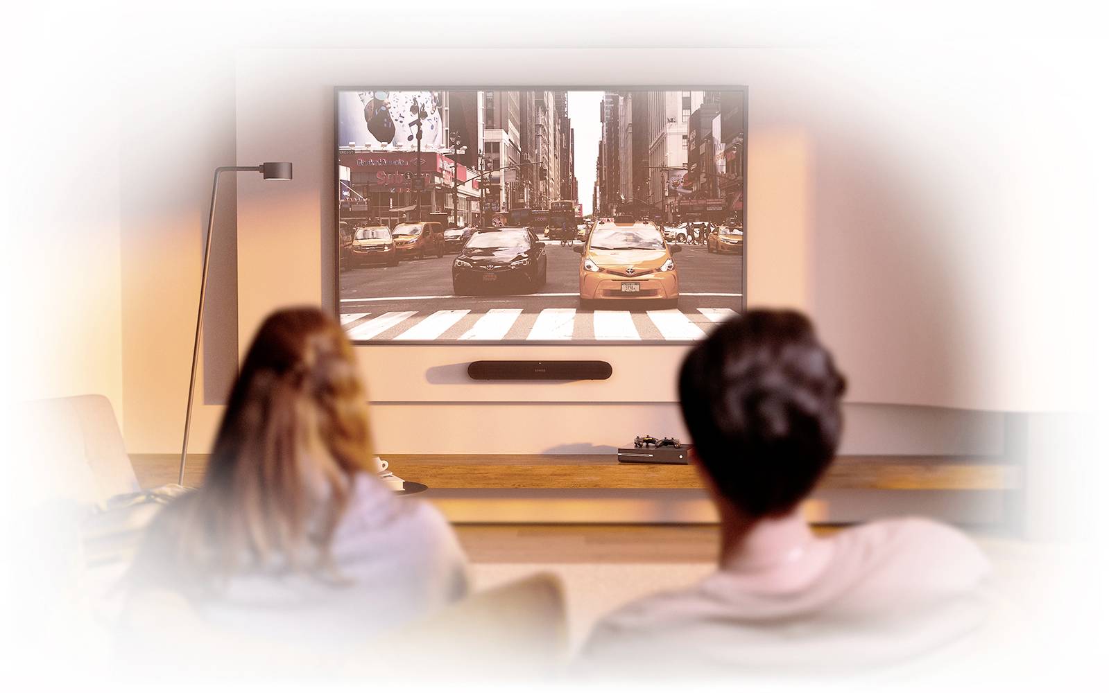 Zwei Personen sitzen in einem Wohnzimmer und schauen einen Film auf einem großen Fernseher, der eine Straßenszene zeigt.