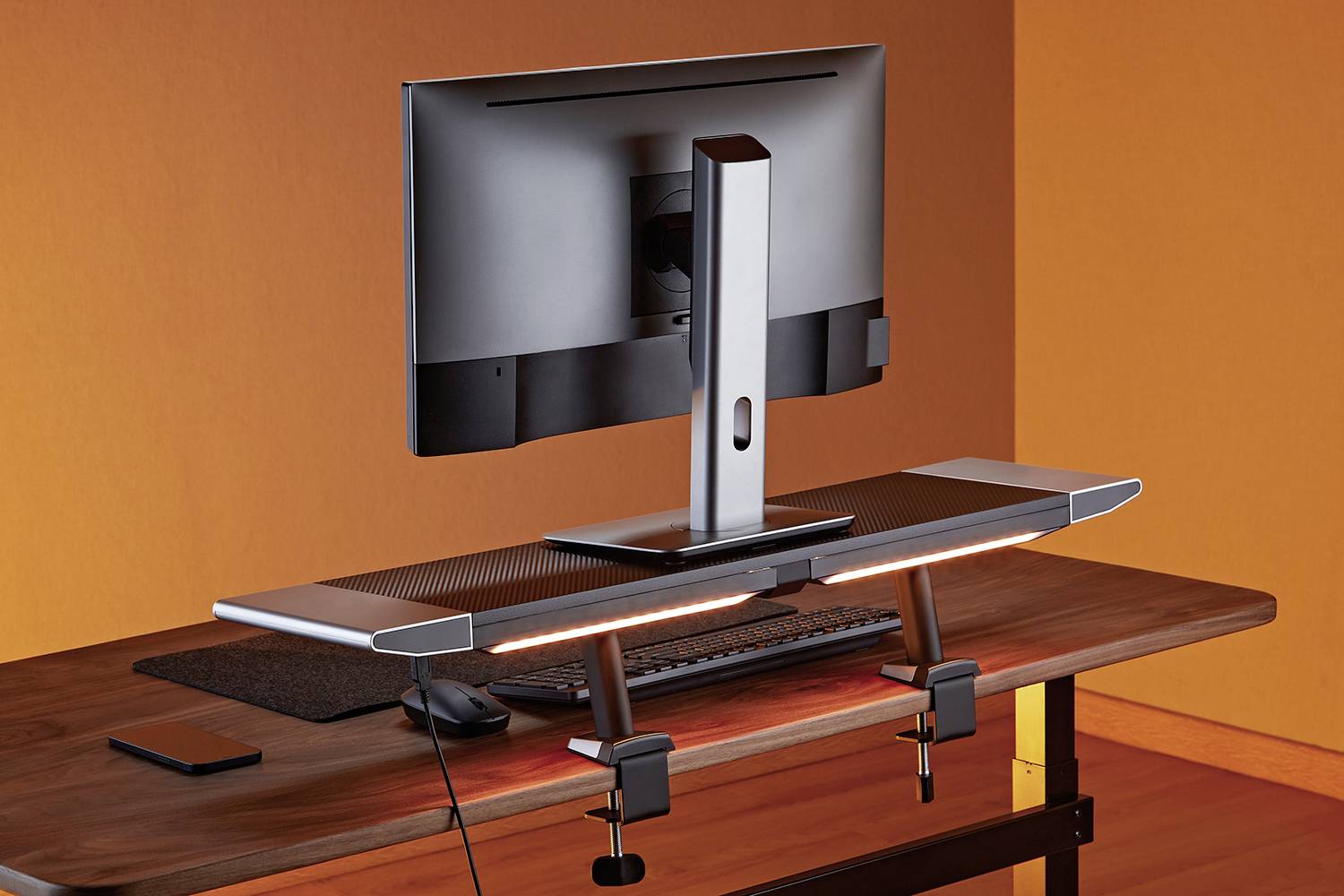 Ein Computerbildschirm und eine Tastatur auf einem Holzschreibtisch. Eine schwarze Lampe mit orangem Licht ist an der Tischkante befestigt.