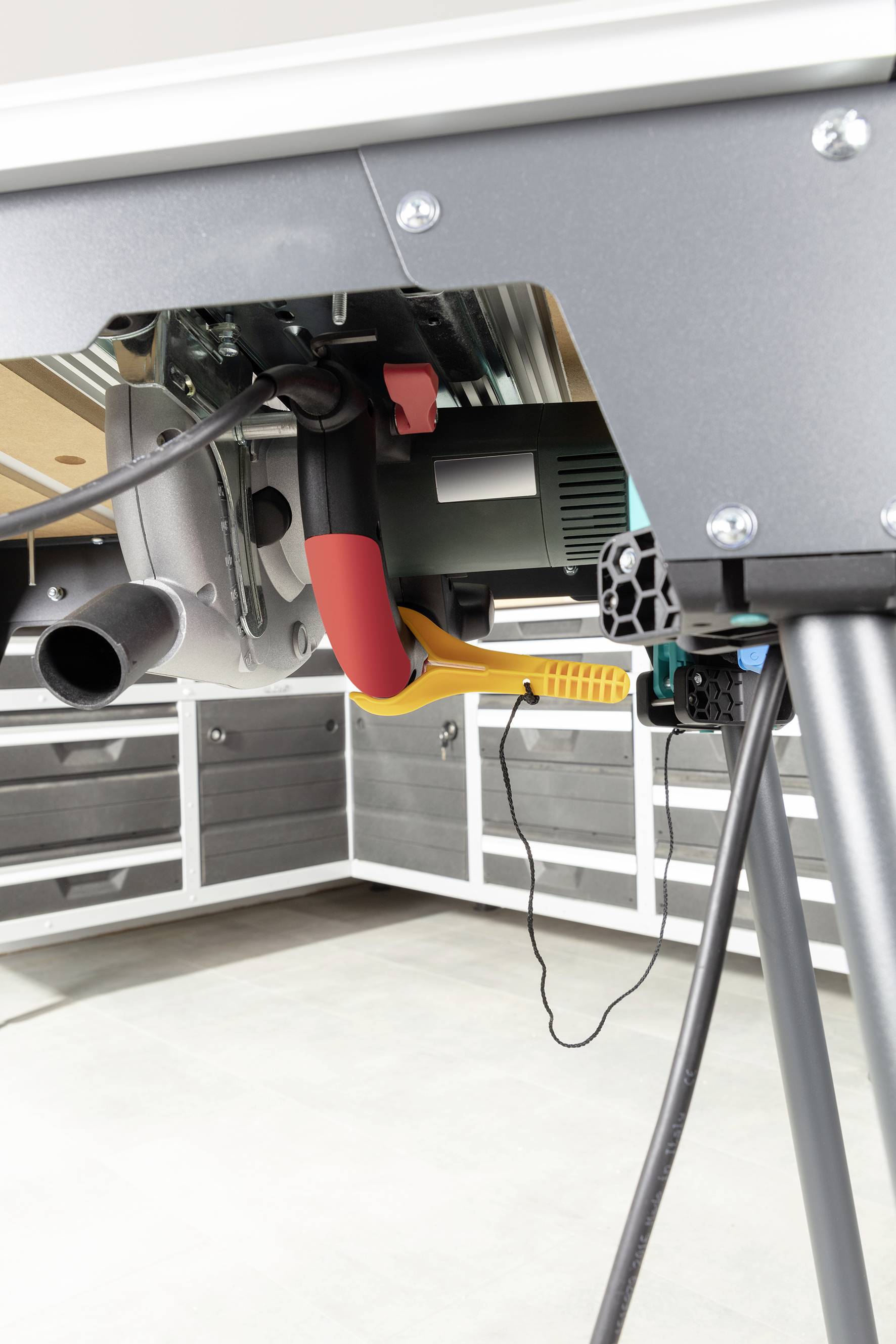 Unteransicht einer Tischsäge mit Staubsaugeranschluss und Stromkabel in einer Werkstatt mit weißen Regalen und grauem Boden.