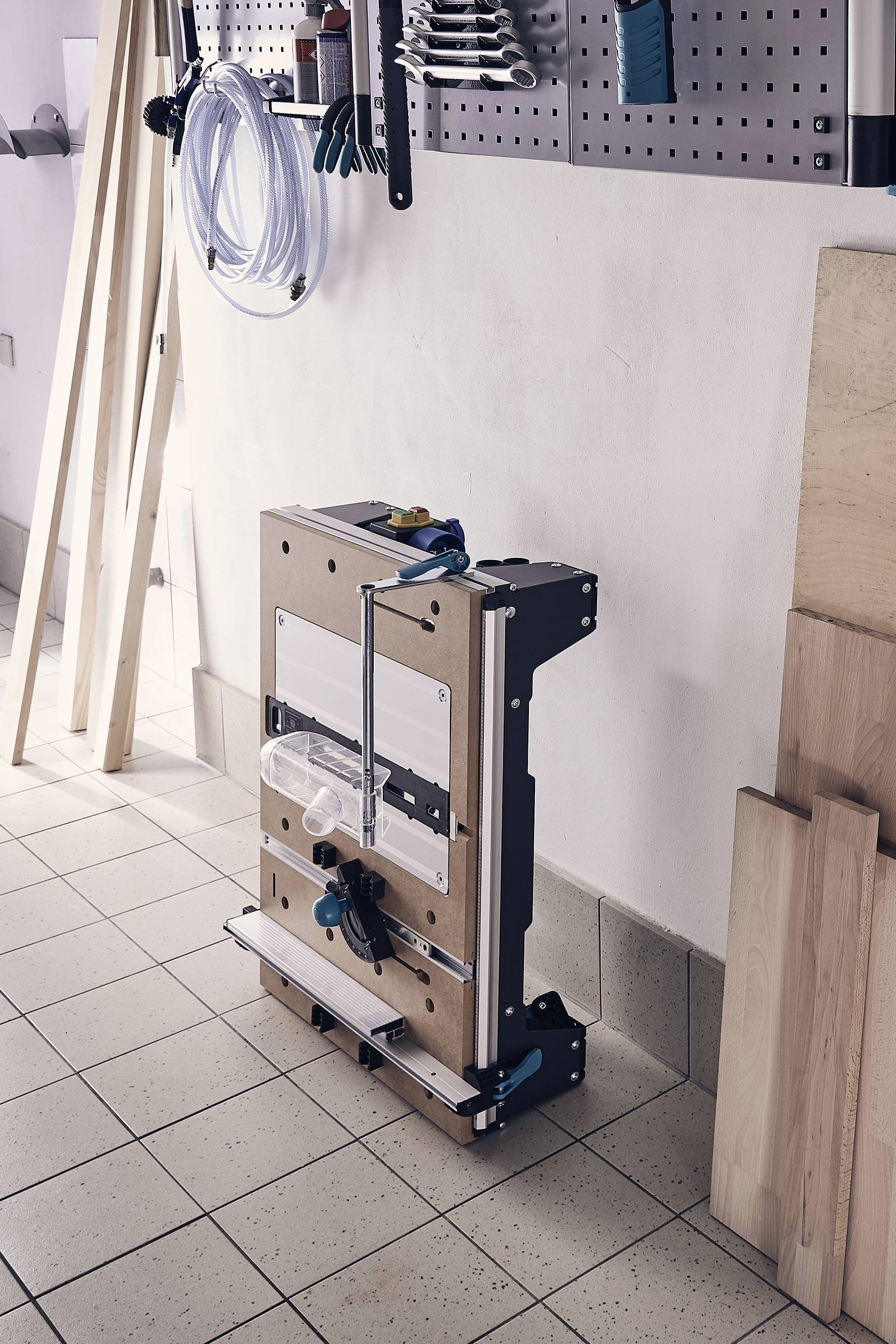 Ein zusammengeklappter Werktisch lehnt an einer weißen Wand in einer Werkstatt. Umliegendes Werkzeug und Holzleisten sind sichtbar.