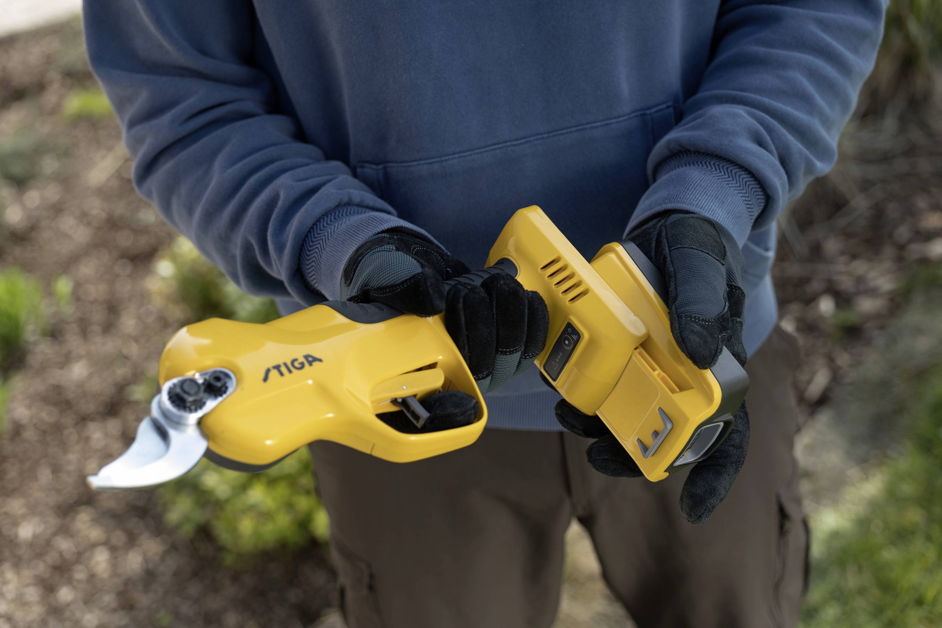 Person hält eine gelbe akkubetriebene Heckenschere. Ihr Akku wird in der anderen Hand gehalten. Gartenhintergrund sichtbar.