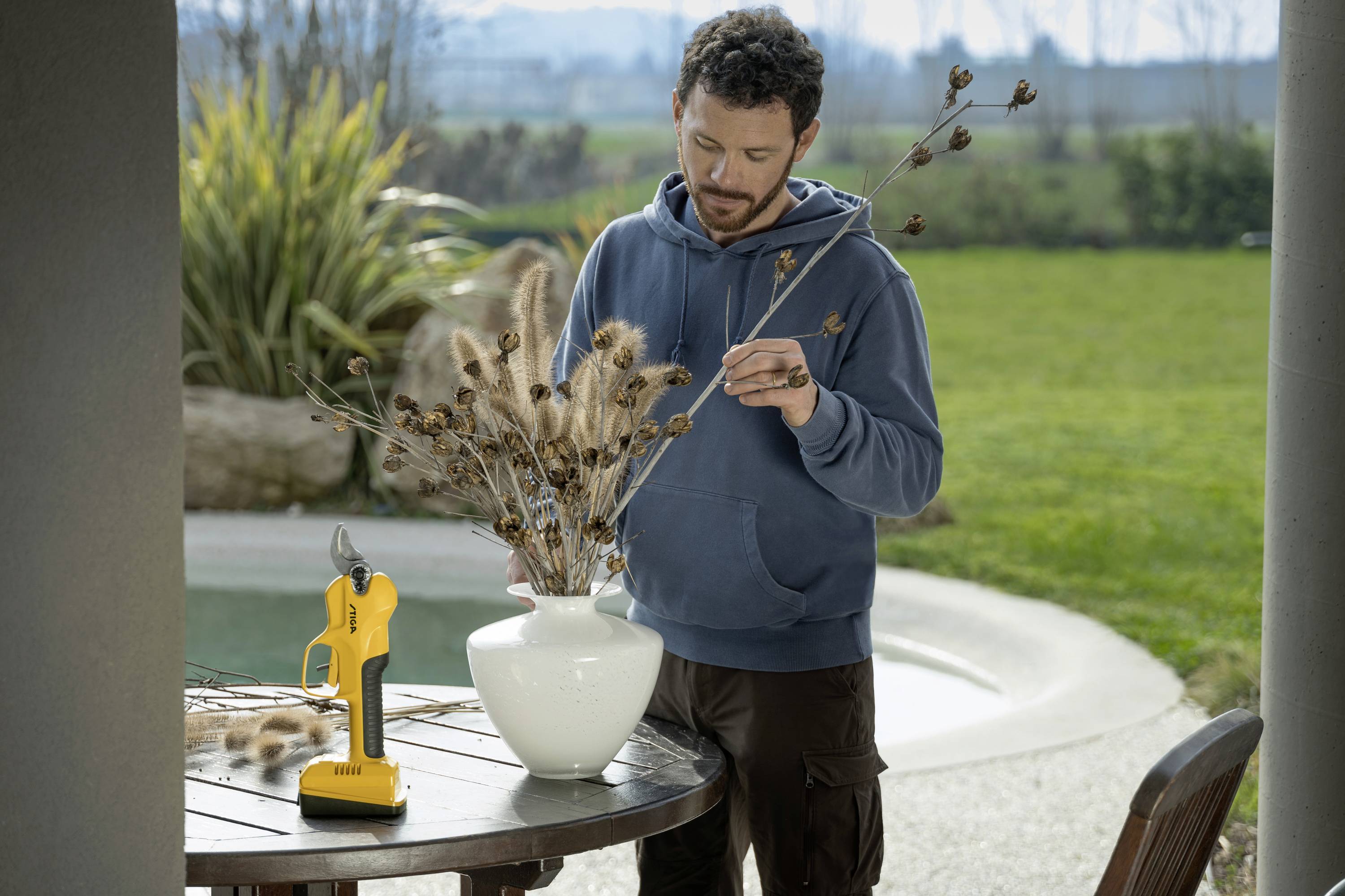 Ein Mann in Freizeitkleidung arrangiert trockene Zweige in einer weißen Vase auf einem Holztisch im Freien. Im Hintergrund Gartenlandschaft.