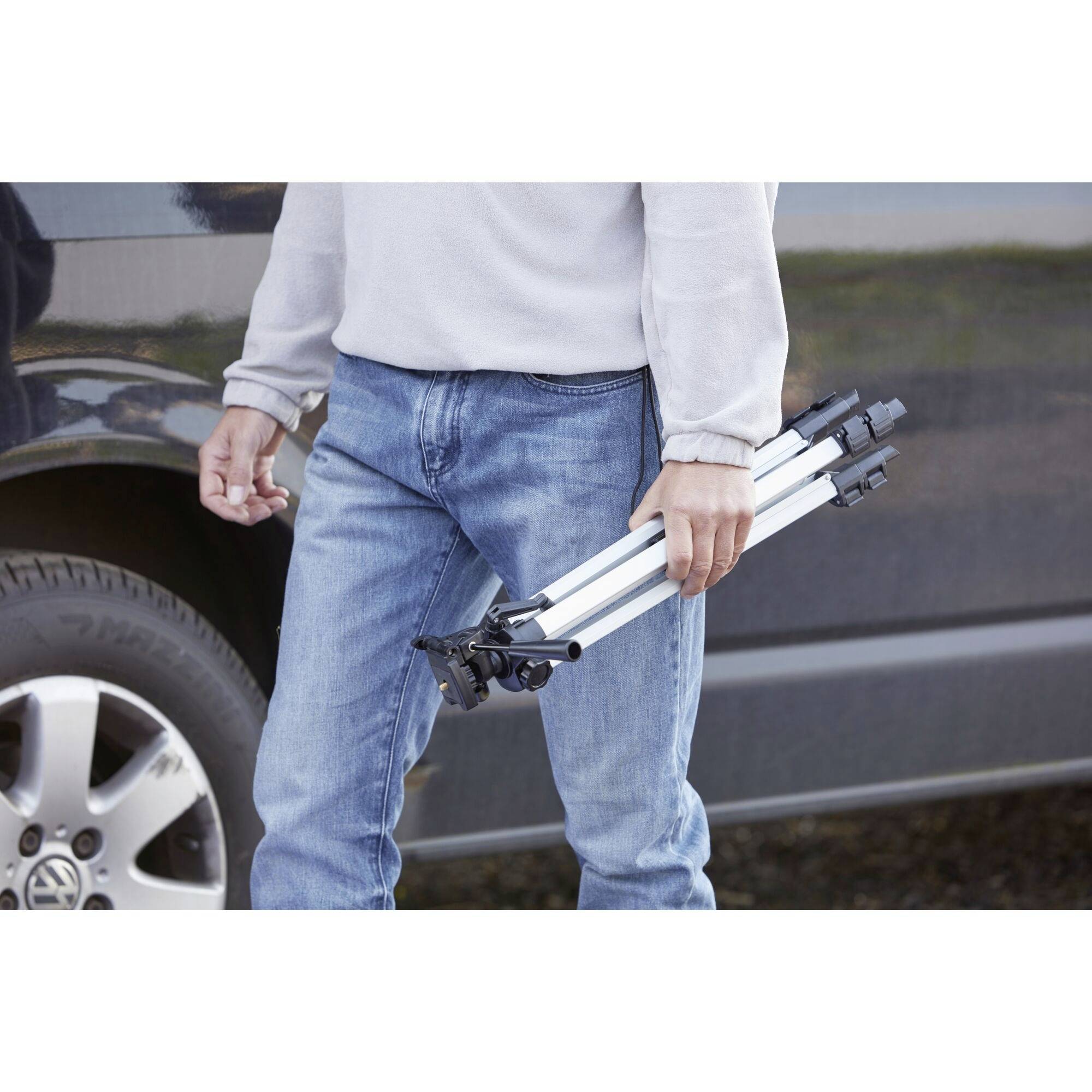 Eine Person hält ein zusammengeklapptes Fotostativ in der Hand. Im Hintergrund ist ein Teil eines Autos zu sehen.
