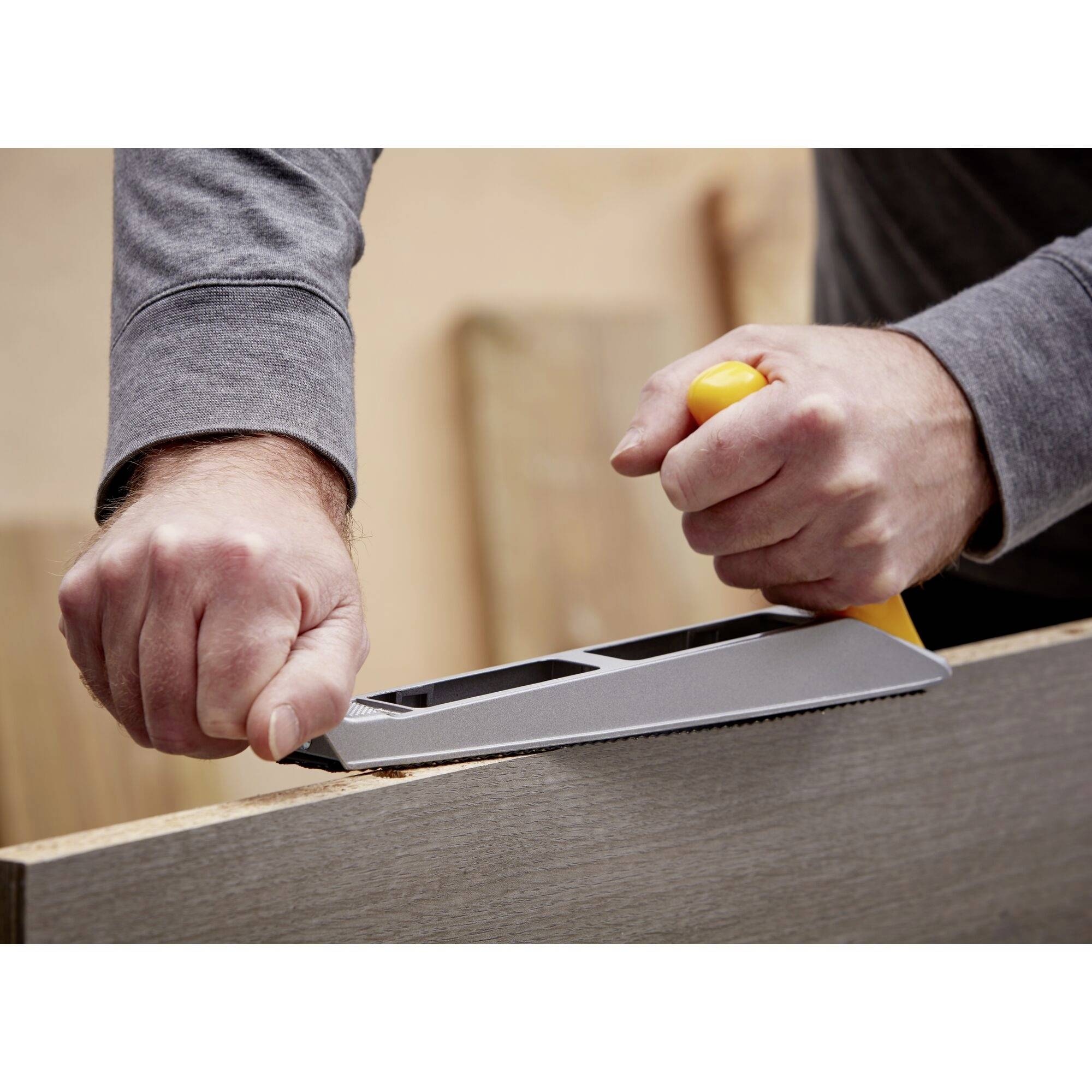 Eine Person hobelt ein Holzstück mit einem Handhobel. Die Hände halten das Werkzeug fest, während Holzspäne entstehen.