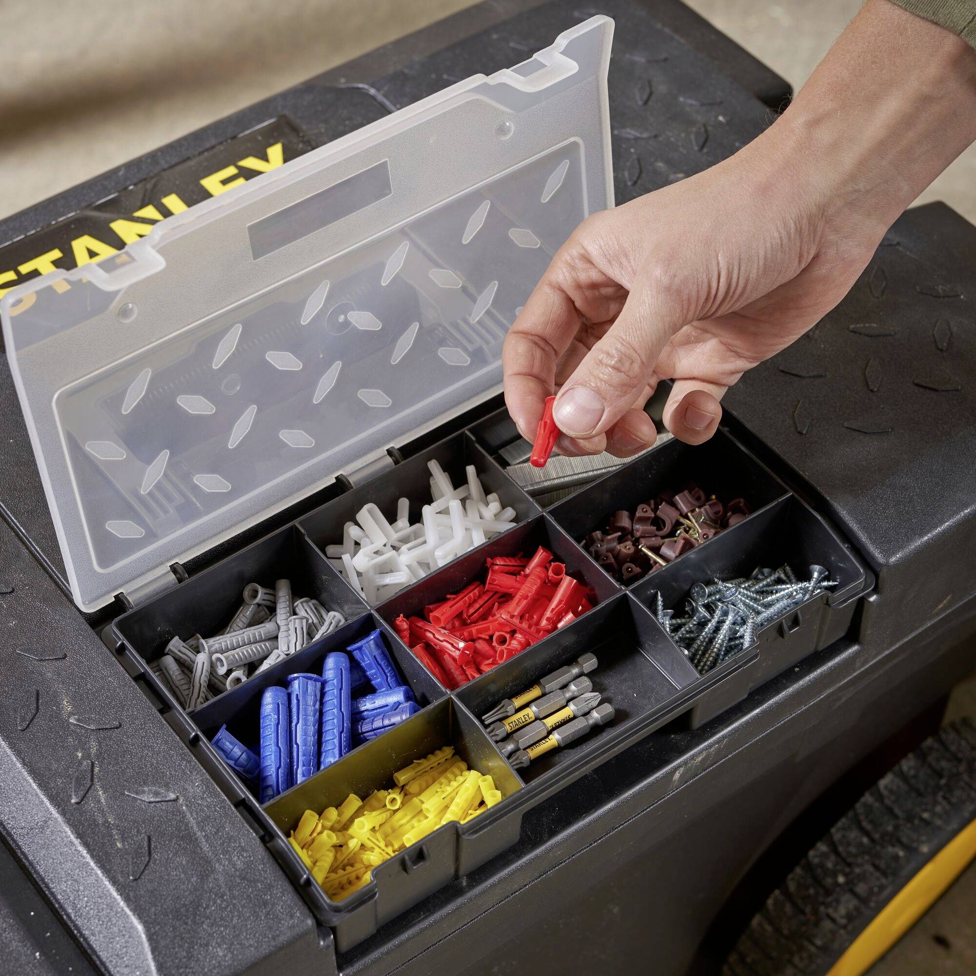 Eine Hand greift nach einem roten Dübel aus einer geöffneten Organizer-Box, die verschiedene Schrauben und Dübel enthält.
