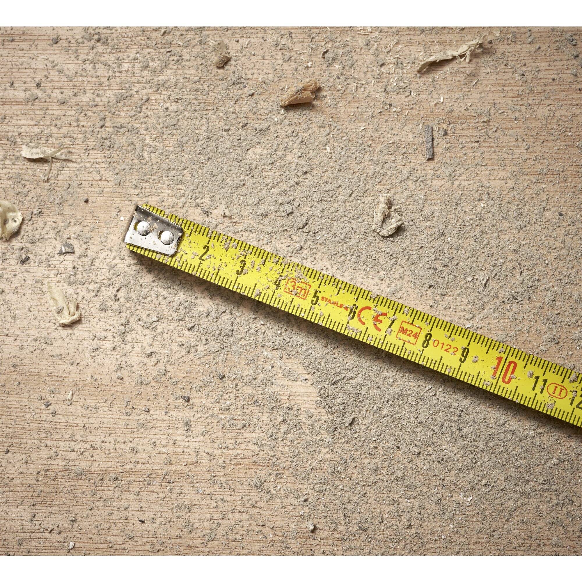 Gelbes Maßband auf staubigem Holzboden mit Holzspänen, zeigt eine handwerkliche oder bauliche Umgebung.