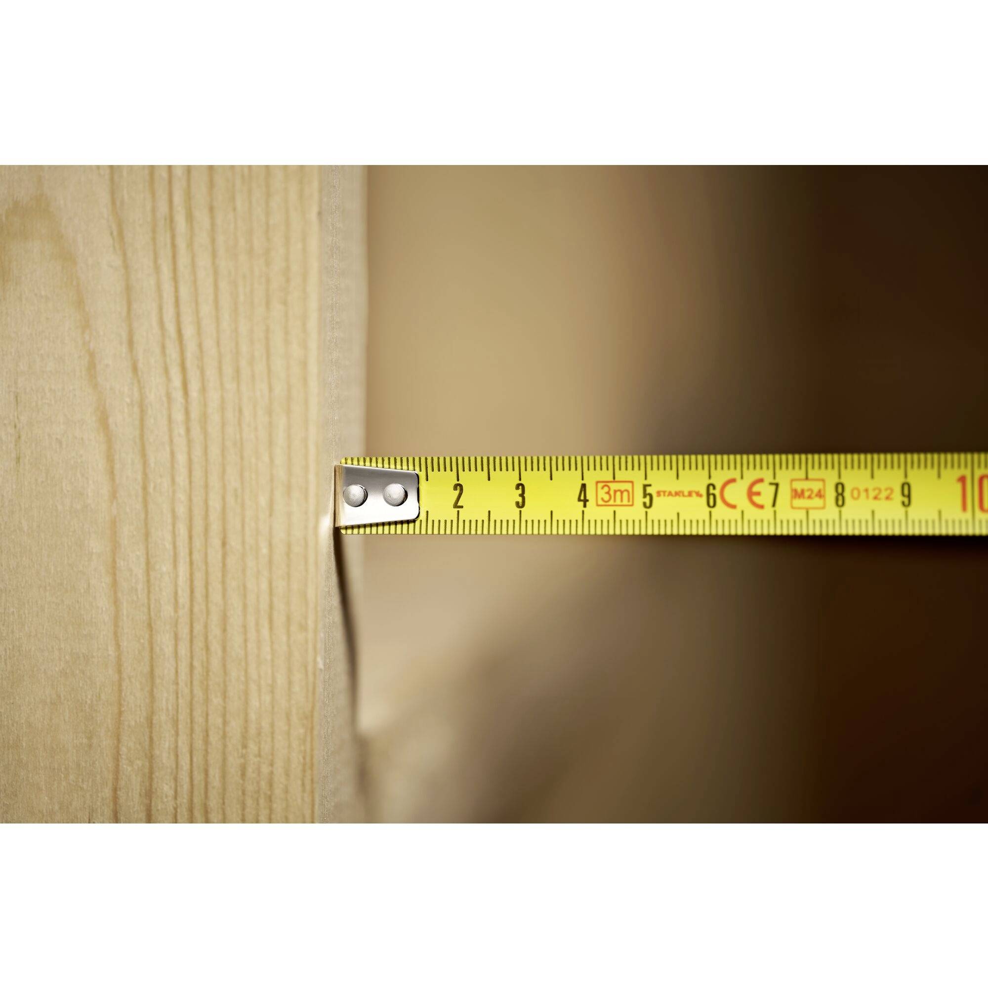 Ein gelbes Maßband misst die Breite eines Holzbretts. Die Markierung liegt bei etwa 4,5 cm.