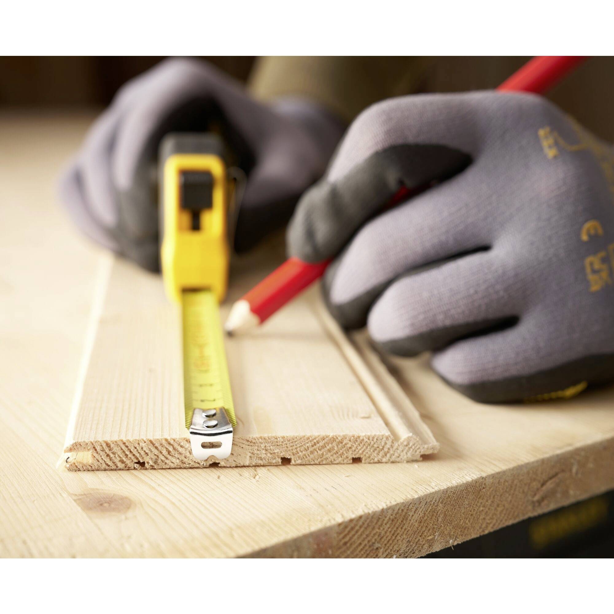 Eine Person mit Handschuhen nutzt ein Maßband und Bleistift, um ein Holzbrett präzise zu markieren.