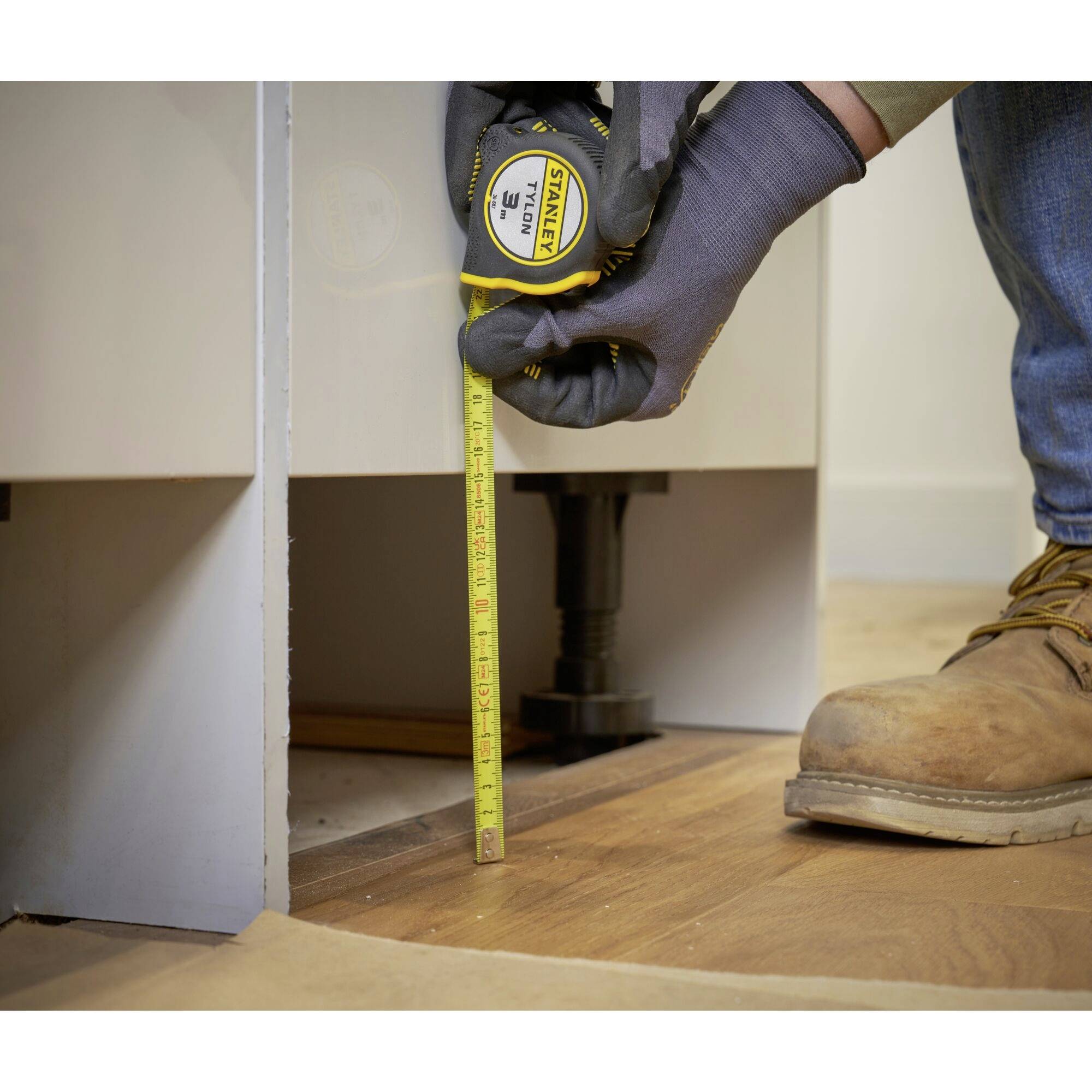 Hand mit Handschuh hält Maßband, misst Höhe zwischen Boden und Möbelunterkante. Person trägt Arbeitshose und Stiefel.