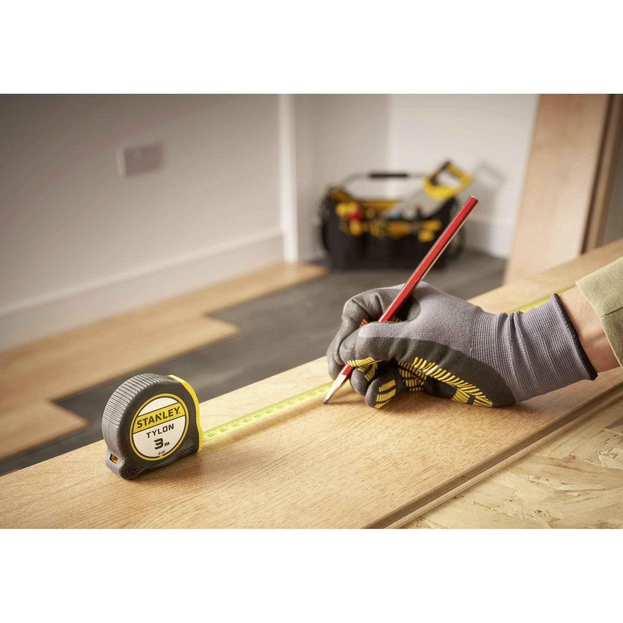 Hand, die mit Handschuh und Bleistift eine Messung auf Holz markiert, neben einem gelben Maßband. Hintergrund zeigt Werkstattumgebung.