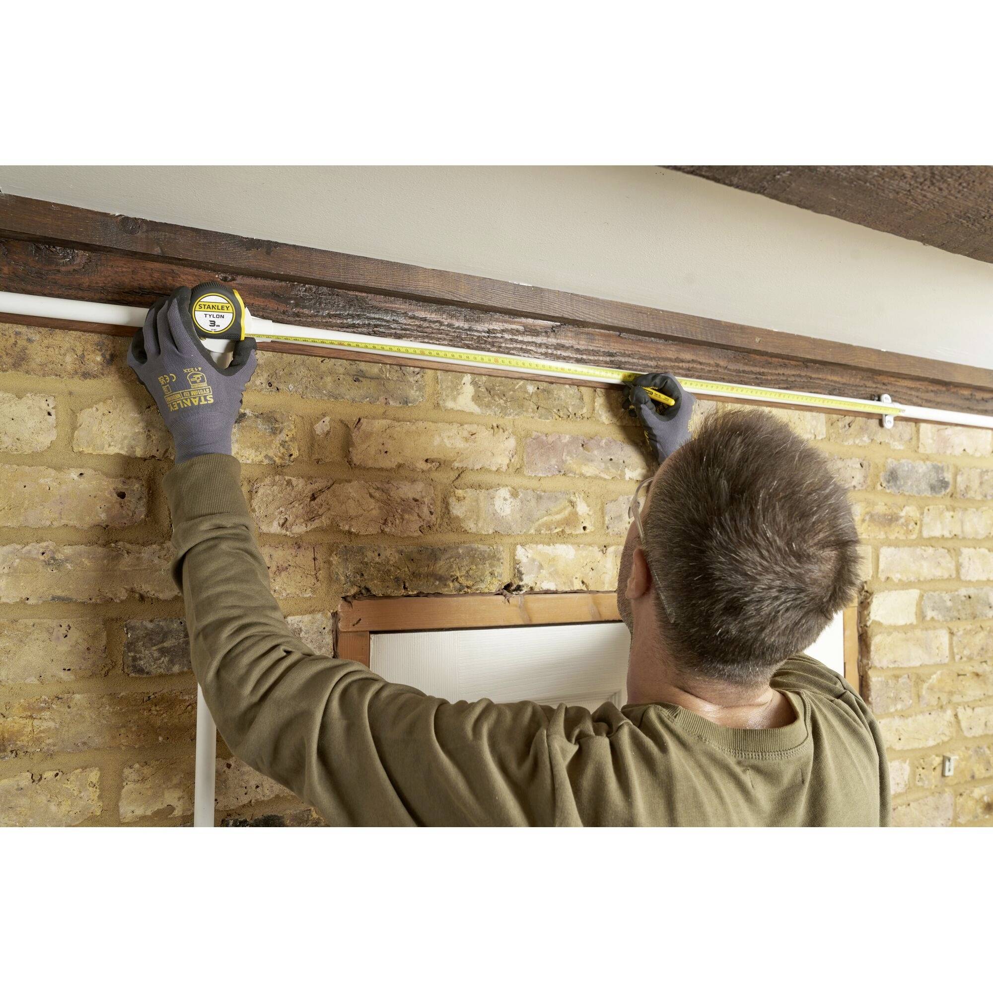 Eine Person misst mit einem Maßband die Breite einer Wand aus Ziegelsteinen. Sie trägt Handschuhe und fokussiert auf die Messung.