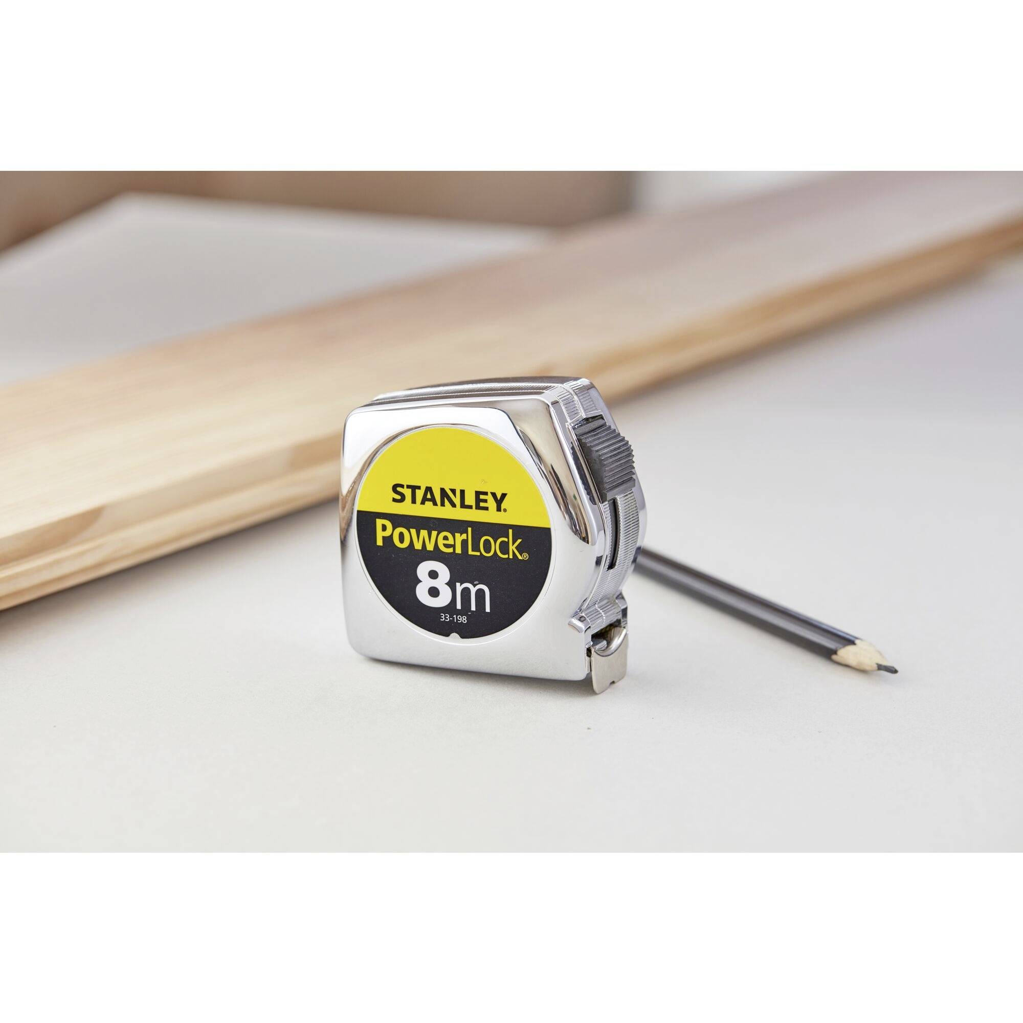 Ein Maßband der Marke Stanley 'PowerLock 8m' liegt auf einem Tisch neben einem Holzbalken und einem Stift.