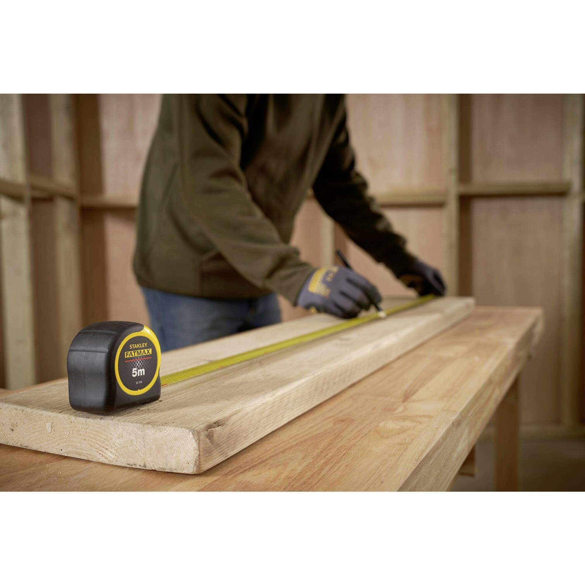 Eine Person misst ein Holzbrett mit einem Maßband in einer Werkstatt. Auf dem Tisch liegt ein 5m-Maßband mit gelbem Gehäuse.