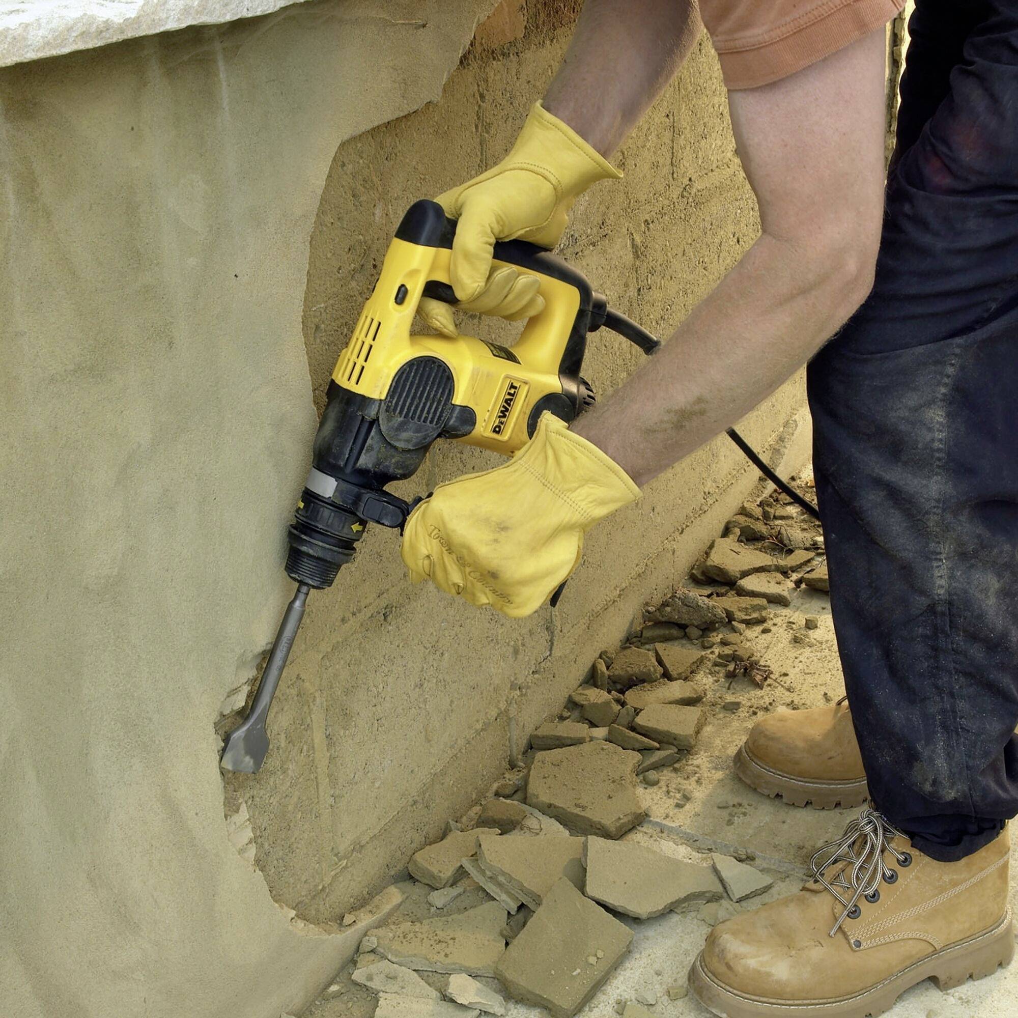 Eine Person benutzt einen gelben Bohrhammer, um eine Wand abzubauen. Sie trägt gelbe Handschuhe und braune Stiefel.