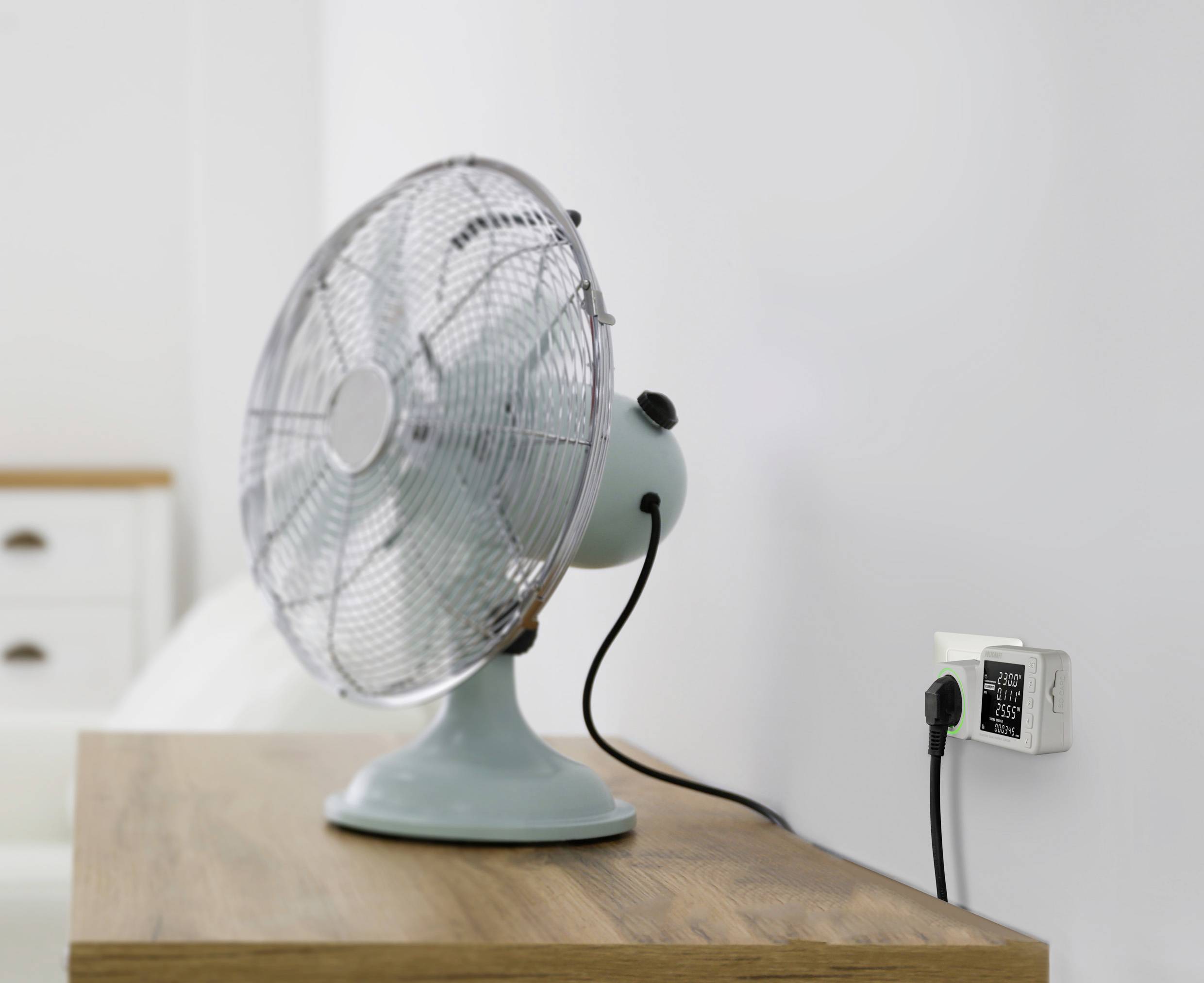 Ein Tischventilator mit blauem Gehäuse steht auf einem Holztisch. Er ist an eine Wandsteckdose angeschlossen. Im Hintergrund ist ein weißes Bett zu sehen.