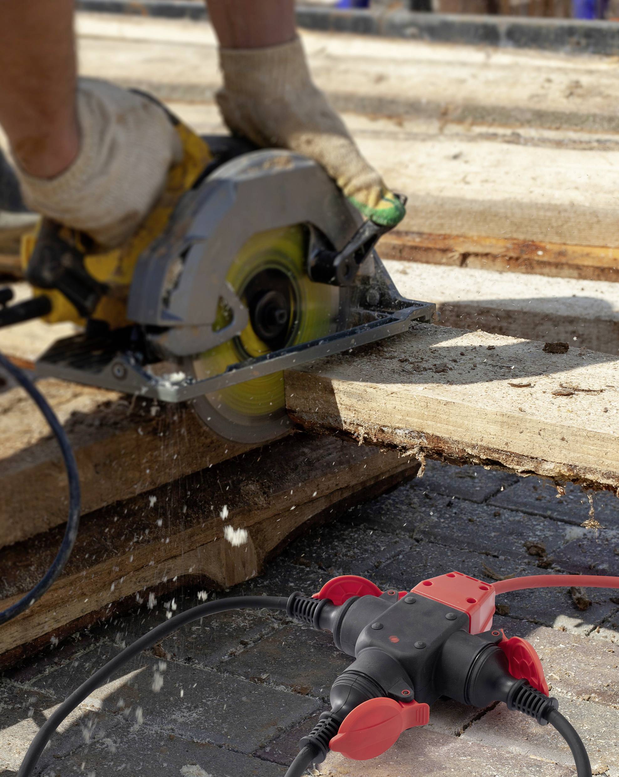 Eine Person arbeitet mit einer Kreissäge, um Holz zu schneiden. Im Vordergrund ist ein Verteiler mit roten Steckern zu sehen.