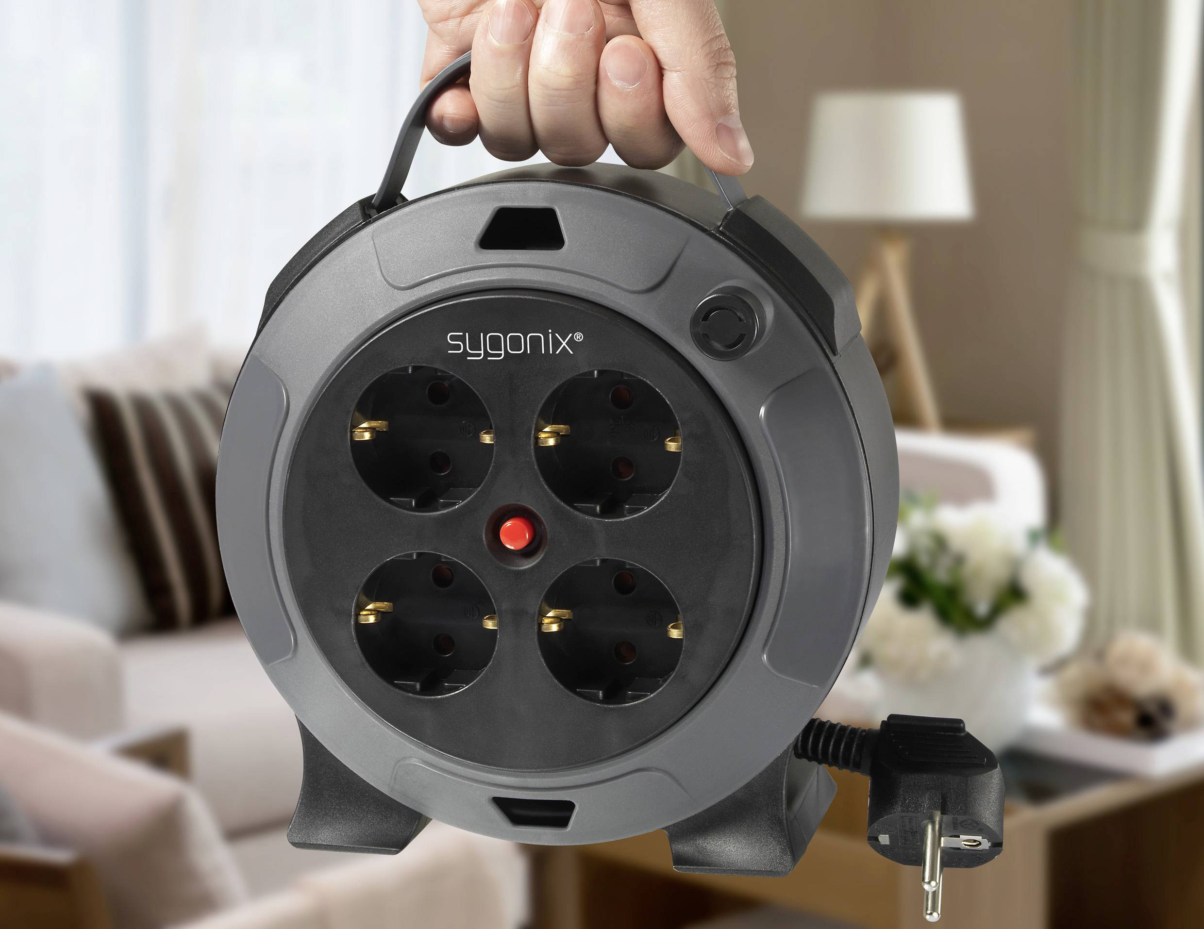 Eine Hand hält einen runden schwarzen Kabeltrommel-Stecker mit mehreren Steckdosen vor einem Wohnzimmerhintergrund.
