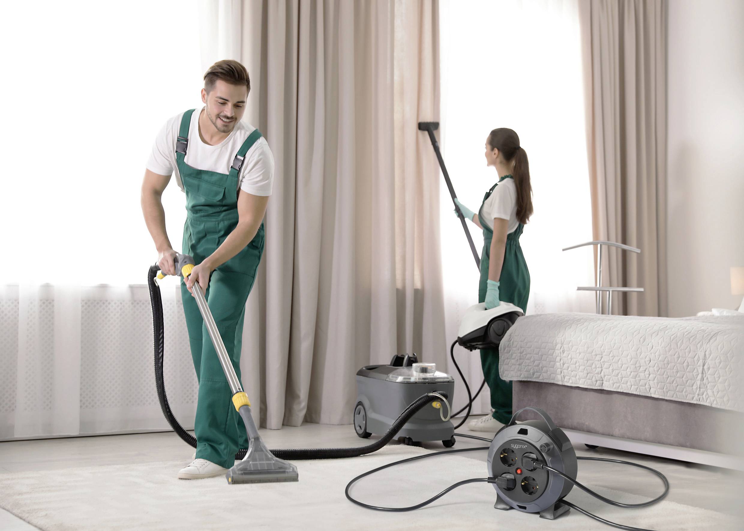 Ein Mann und eine Frau in grünen Overalls reinigen einen Teppich in einem Schlafzimmer mit Dampfreinigern.