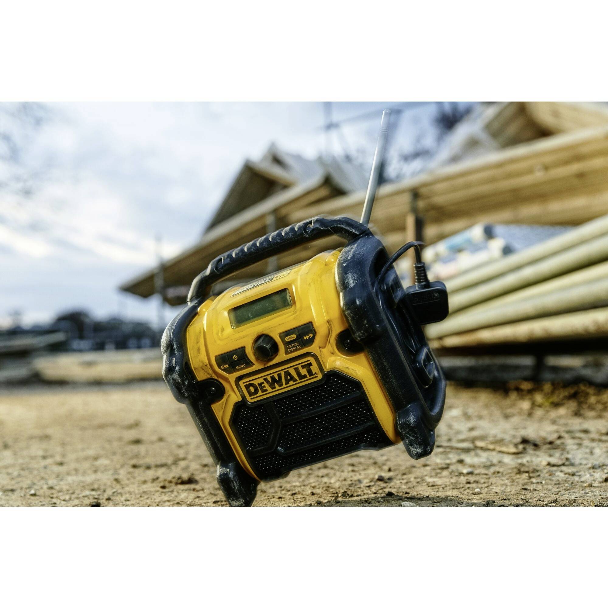 Baustellenradio in Gelb und Schwarz liegt auf sandigem Boden, Hintergrund zeigt unscharfe Holzstapel.