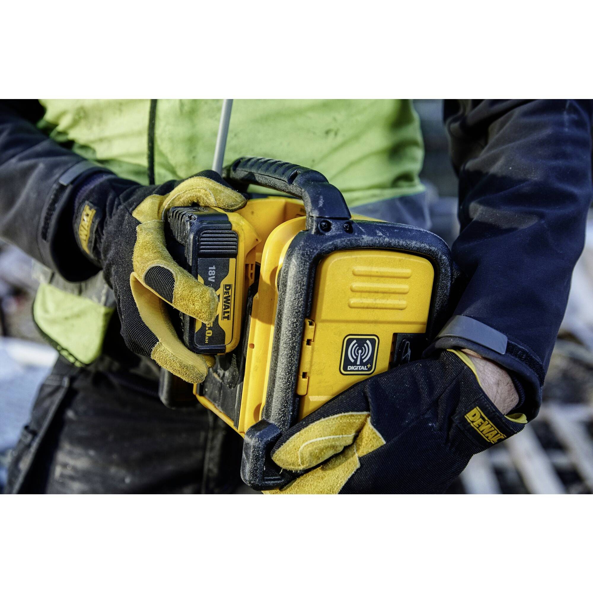 Eine Person hält ein gelbes tragbares Baustellenradio mit schwarzen Elementen. Das Radio hat einen auffälligen Lautsprechergitter.