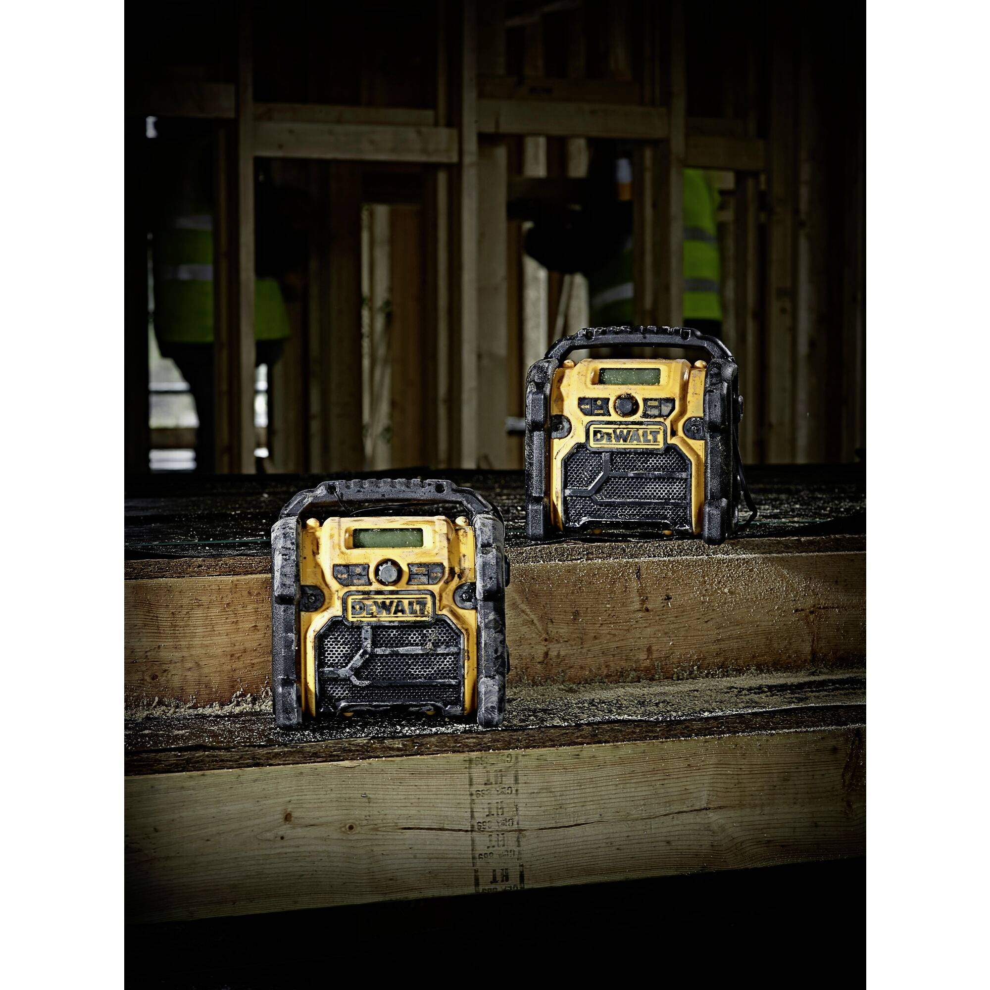 Zwei gelb-schwarze tragbare Radios stehen auf Holz in einem Rohbau. Im Hintergrund sind unscharf Menschen in Baukleidung erkennbar.