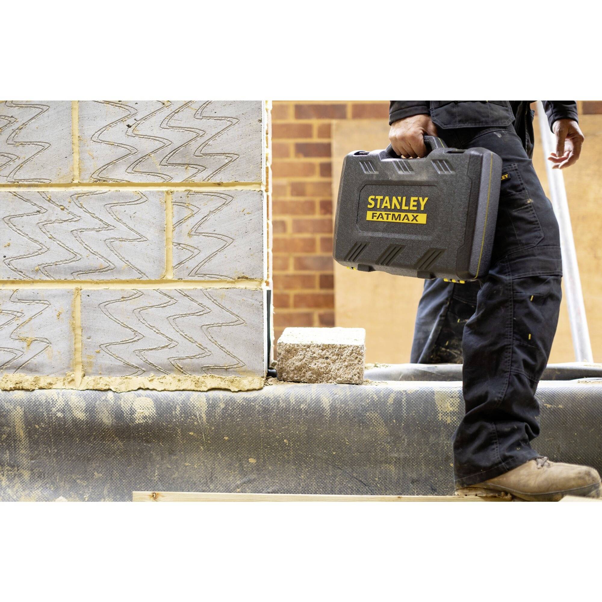 Eine Person trägt einen 'Stanley Fatmax' Werkzeugkoffer entlang einer Baustelle, an einer Wand aus grauen Betonblöcken vorbei.