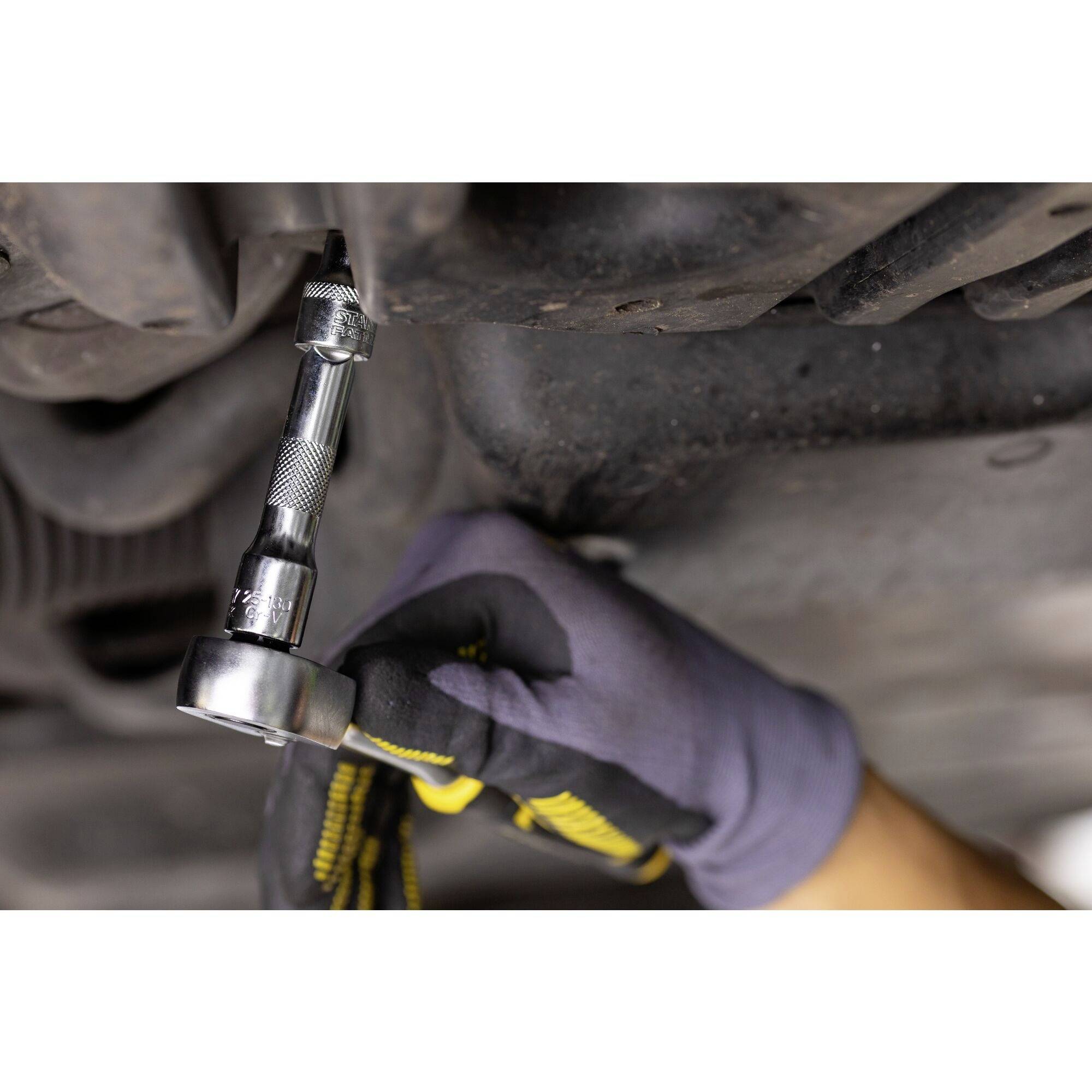 Hand mit Handschuh verwendet Ratsche, um Schraube an Fahrzeugunterseite zu lösen. Mechanikerarbeit an Auto.