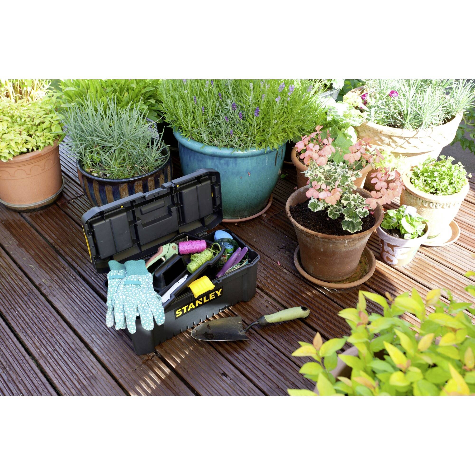 Gartenarbeitsplatz mit Werkzeugkasten, Handschuhen und Schaufel neben Pflanzen in Töpfen auf einer Holzterrasse.