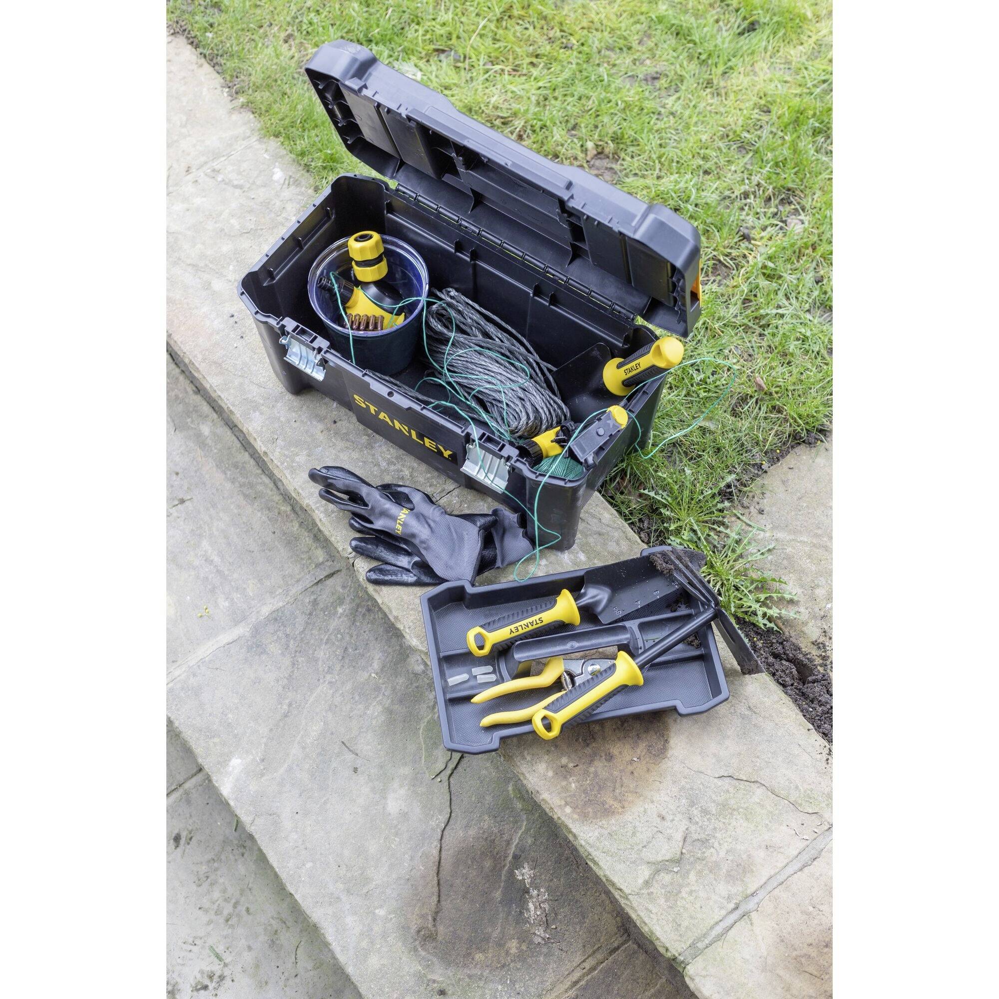 Offene Werkzeugkiste auf einem Gehweg, enthält Handschuhe, Drahtspule und gelb-schwarze Werkzeuge. Gras im Hintergrund.