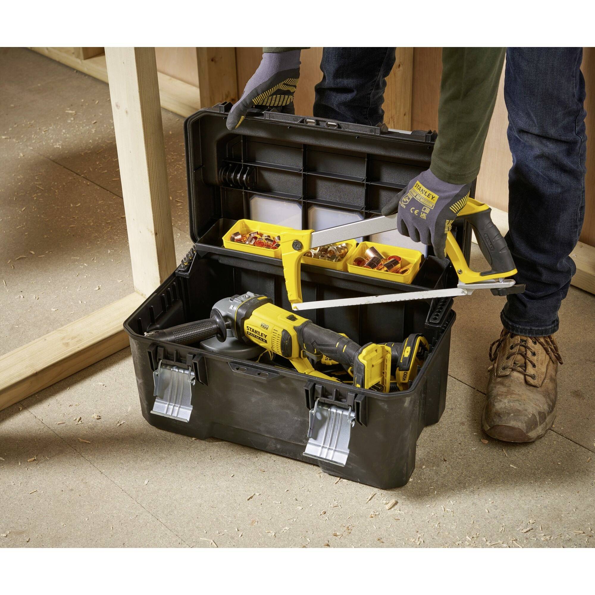 Ein Arbeiter öffnet einen Werkzeugkasten mit verschiedenen Werkzeugen wie einer Handsäge und einer Bohrmaschine. Im Hintergrund ist eine Holzrahmenkonstruktion zu sehen.