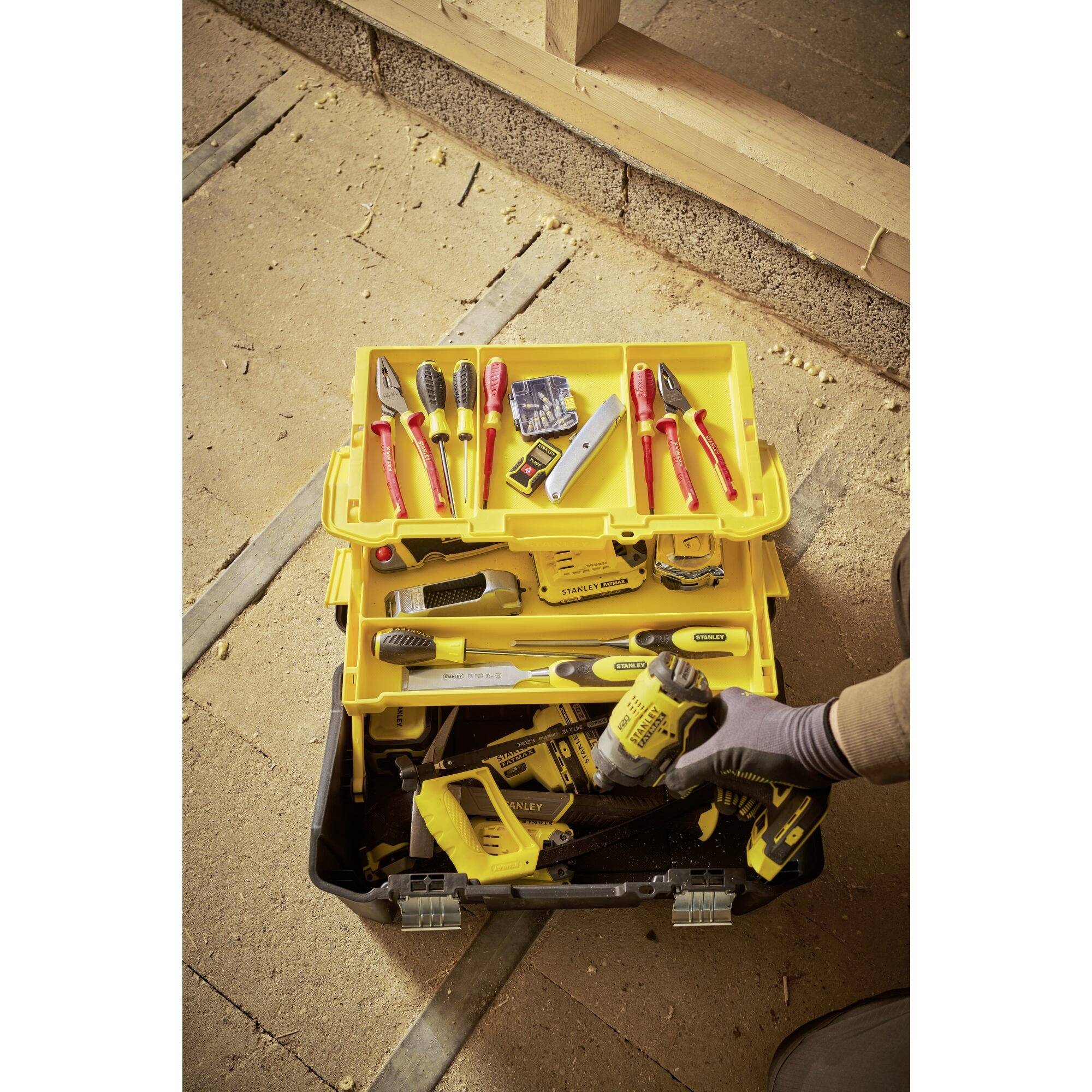 Werkzeugkasten auf Baustelle geöffnet, enthält diverse Werkzeuge wie Schraubendreher, Zangen und Wasserwaage. Ein Handschuh hält eine Bohrmaschine.