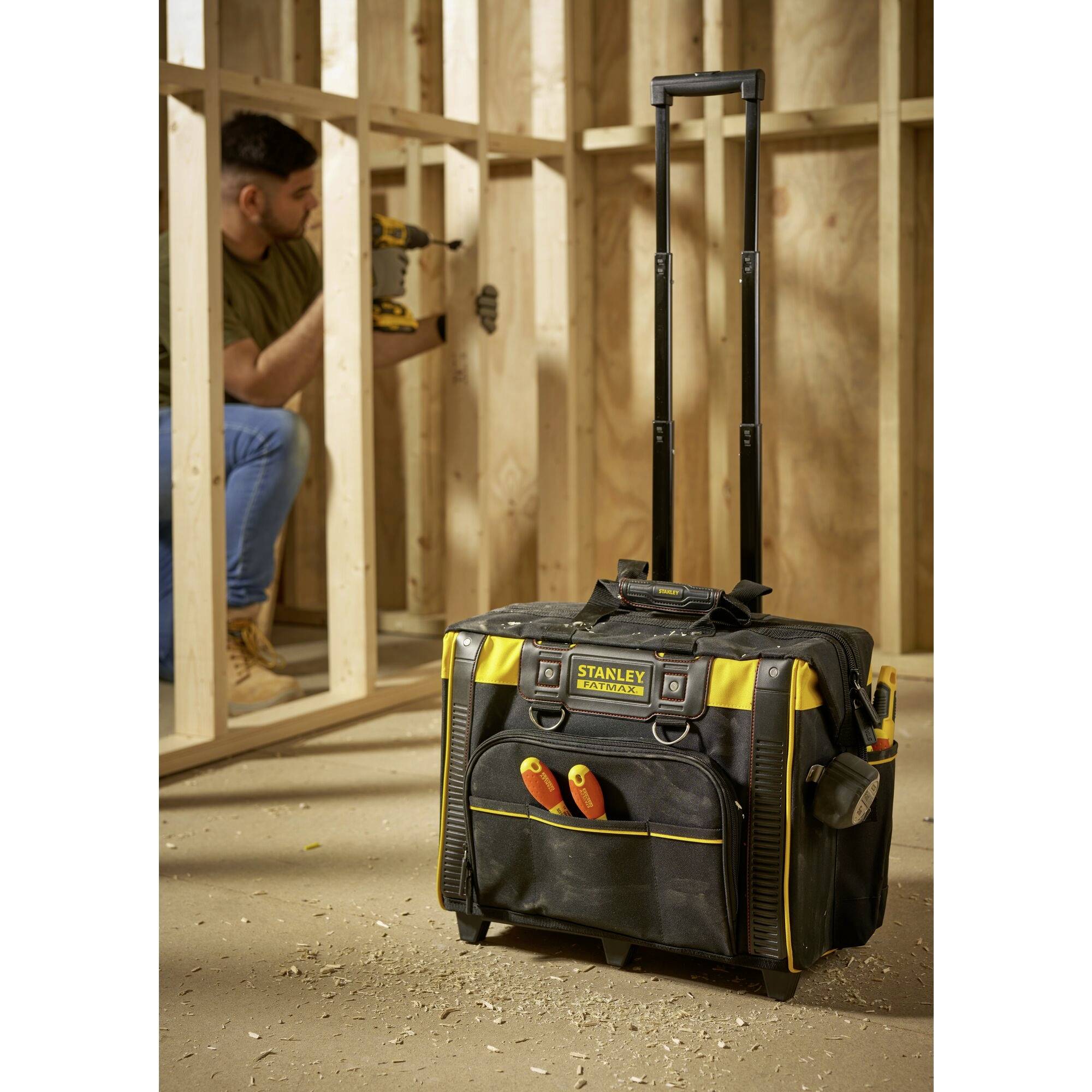 Eine schwarze Werkzeugtasche mit gelben Akzenten und Teleskopgriff steht auf dem Boden einer Holzrahmen-Baustelle; ein Mann nutzt eine Bohrmaschine.