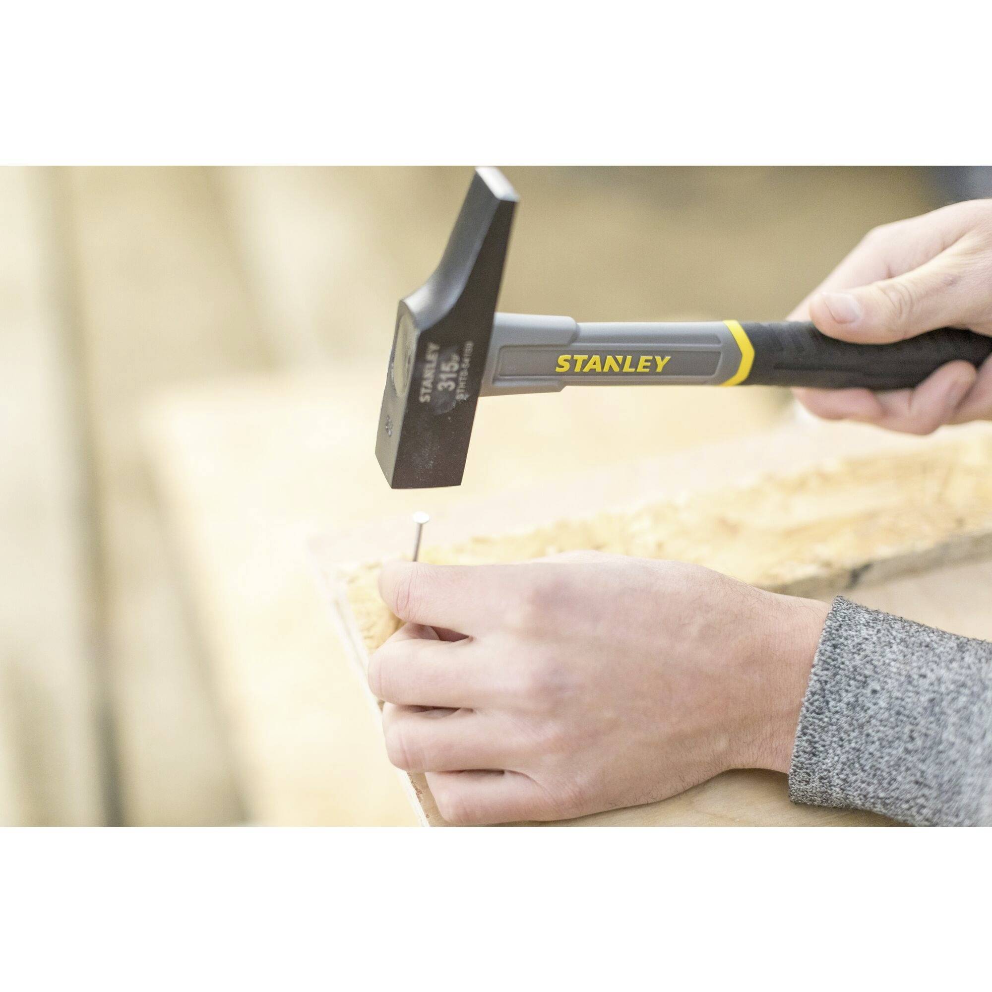 Eine Person hält einen Hammer in der Hand und schlägt einen Nagel in ein Holzbrett. Der Hammer hat die Aufschrift 'Stanley'.