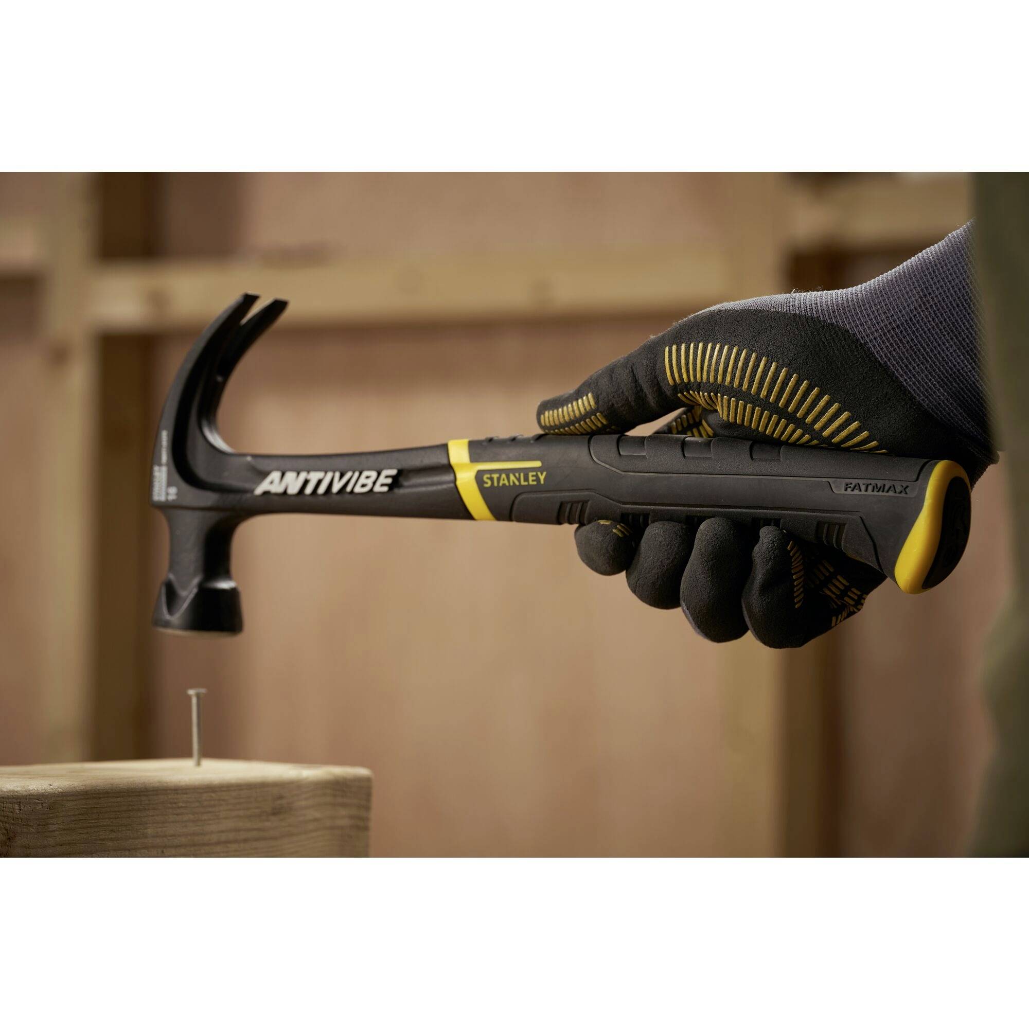 Eine Hand in Arbeitshandschuh hält einen Hammer mit der Aufschrift 'ANTIVIBE STANLEY', bereit, einen Nagel in Holz zu schlagen.