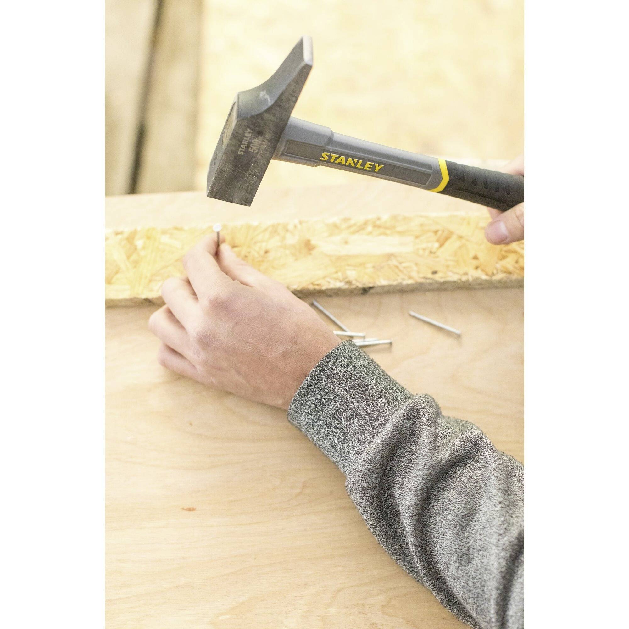 Eine Person hämmert auf einem Holztisch und hält einen Hammer mit der Aufschrift 'STANLEY'. Drei Nägel liegen auf dem Tisch bereit.