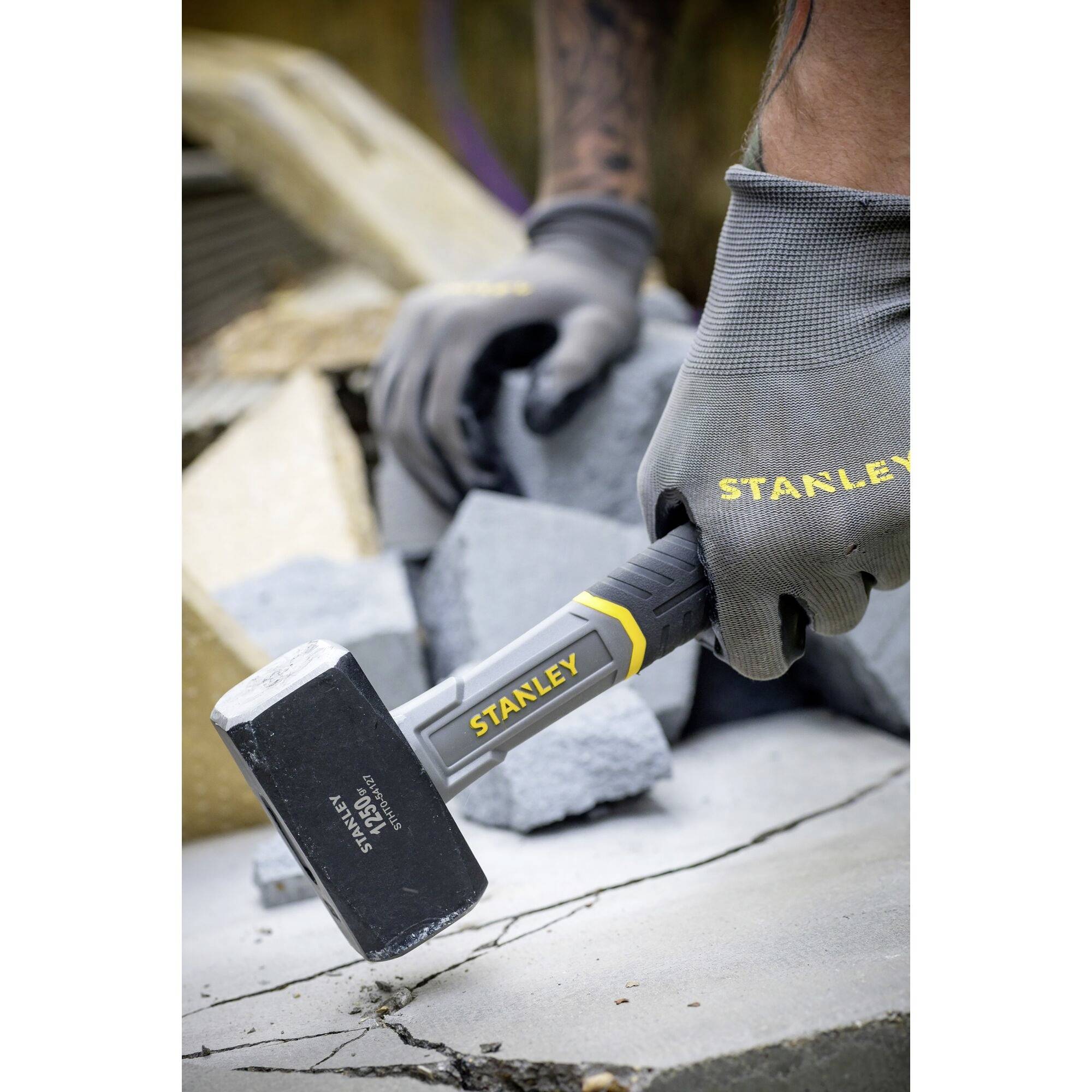Hand in grauem Handschuh hält einen Stanley Hammer, der auf Betonplatte trifft. Im Hintergrund zerbrochene Betonstücke sichtbar.