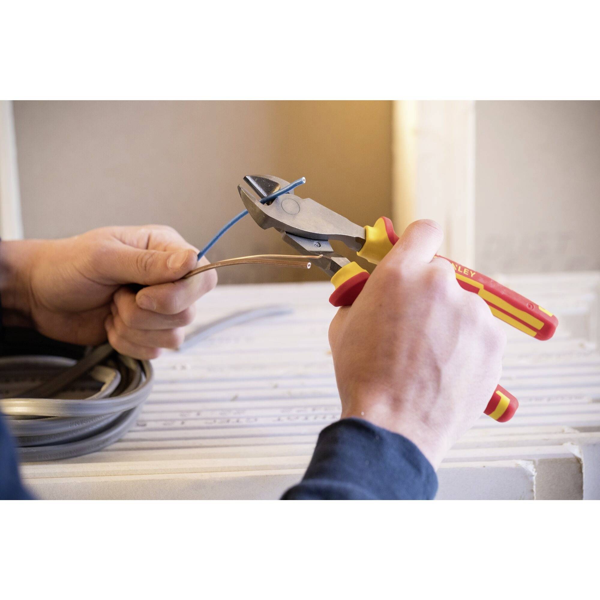 Ein Handwerker schneidet mit isolierten Zangen ein blaues Kabel. Er trägt dunkle Kleidung und arbeitet in einem Raum mit Holzstruktur.