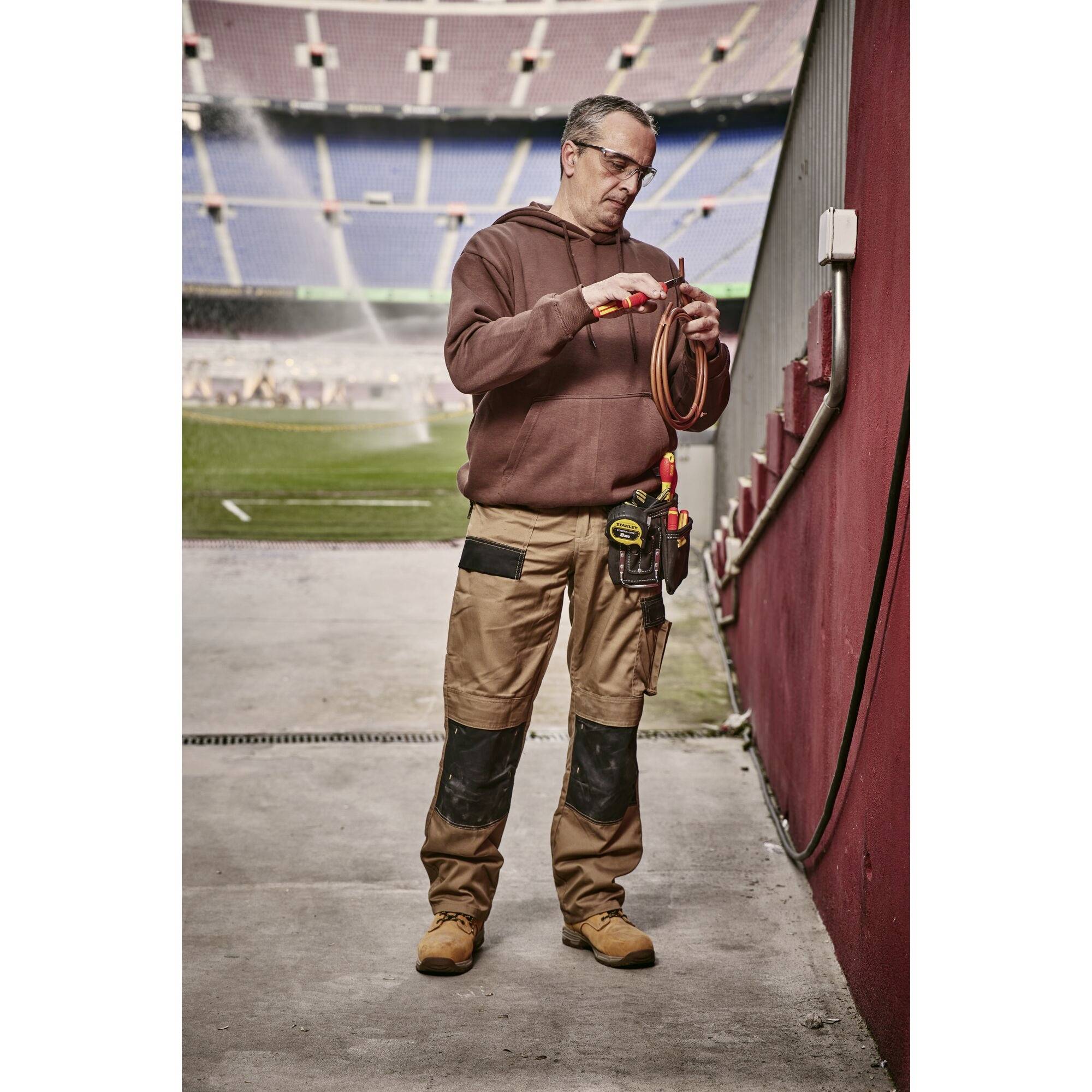 Ein Handwerker mit Werkzeugtasche steht in einem Stadion und ordnet Kabel. Im Hintergrund ist das Spielfeld sichtbar.