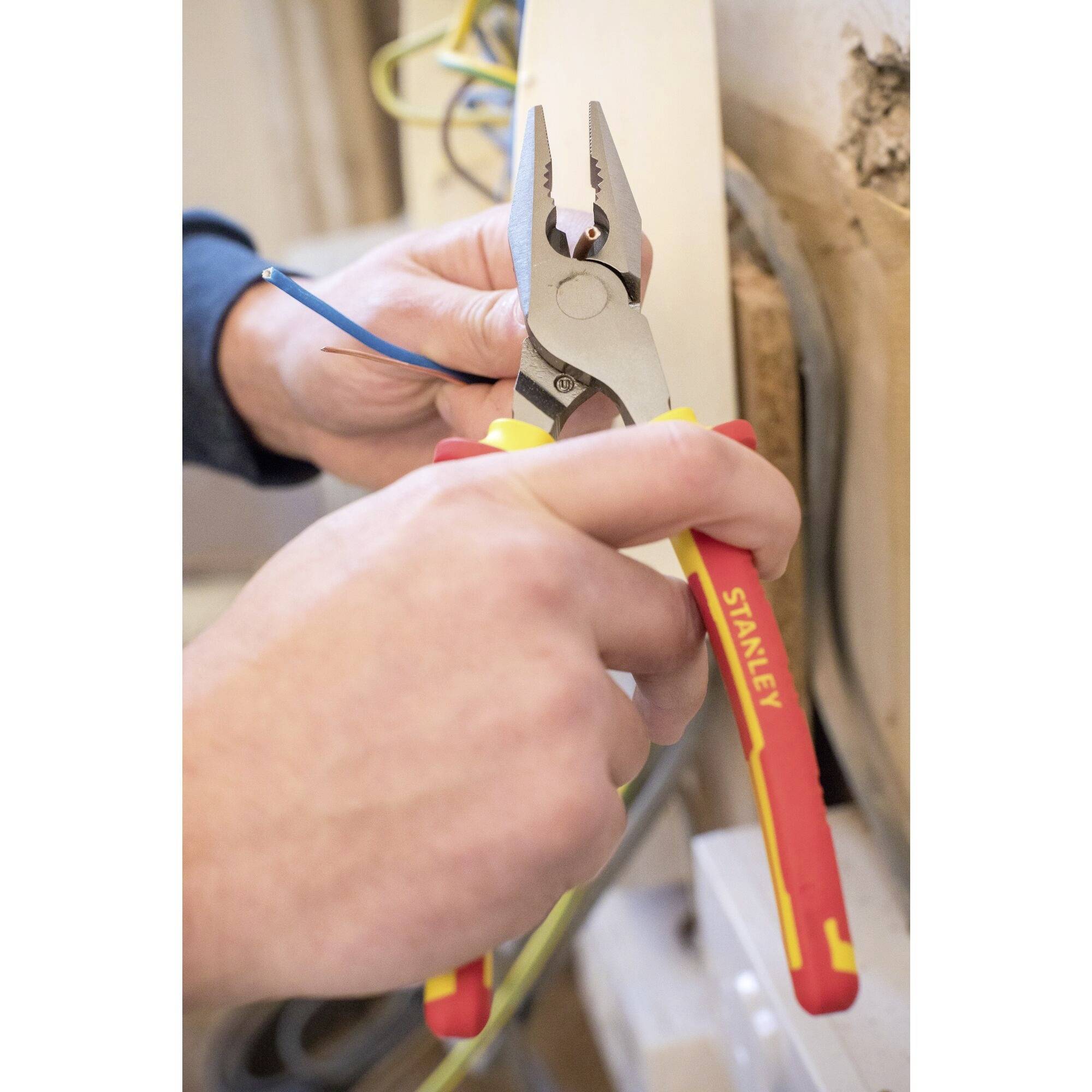 Eine Person benutzt eine Zange, um ein blaues Kabel zu bearbeiten. Sie arbeitet an einem Holzbalken, vermutlich bei Elektroinstallationsarbeiten.