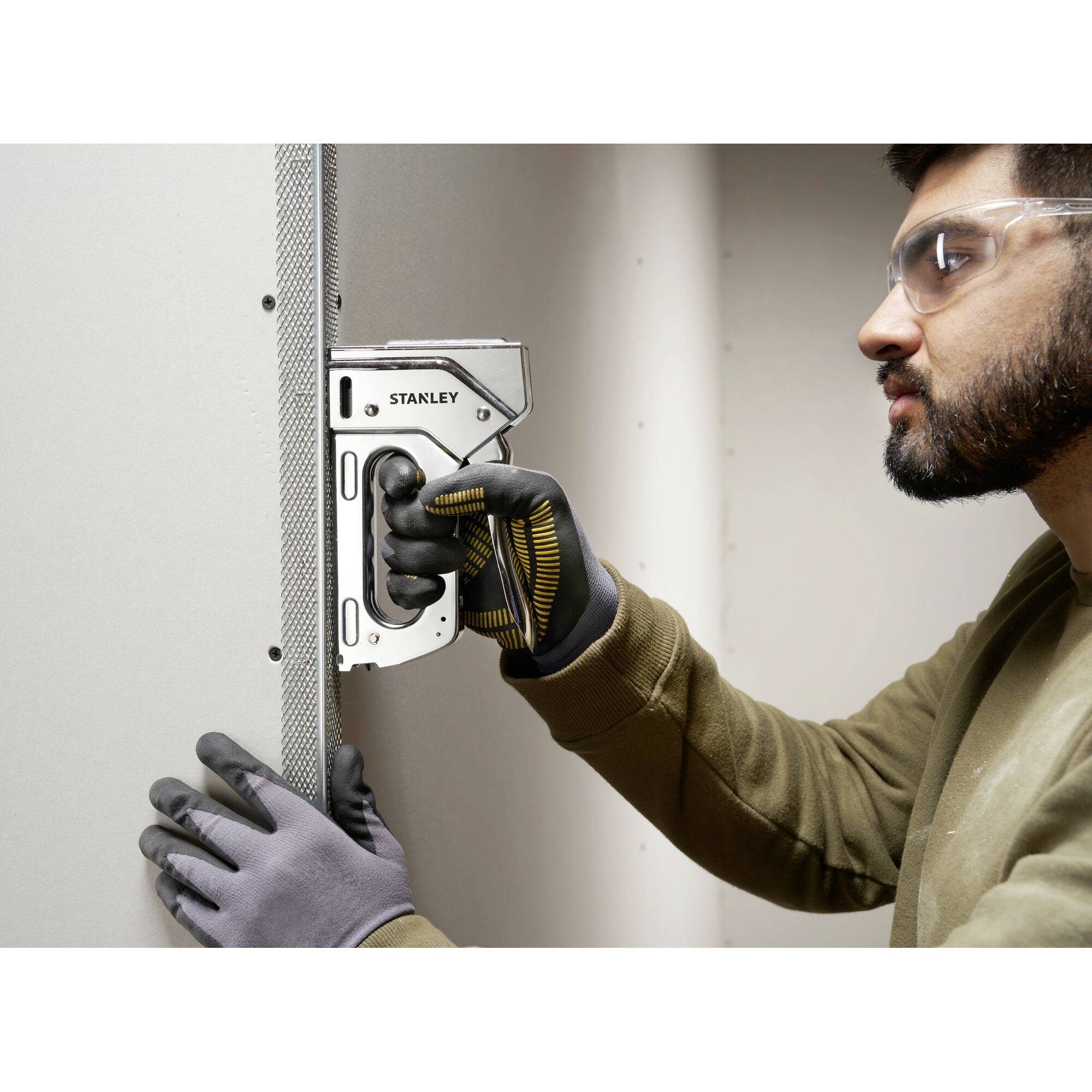 Person mit Schutzbrille und Handschuhen befestigt Gitter auf Rigipswand mit Tacker, Fokus auf präziser Handhabung des Werkzeugs.