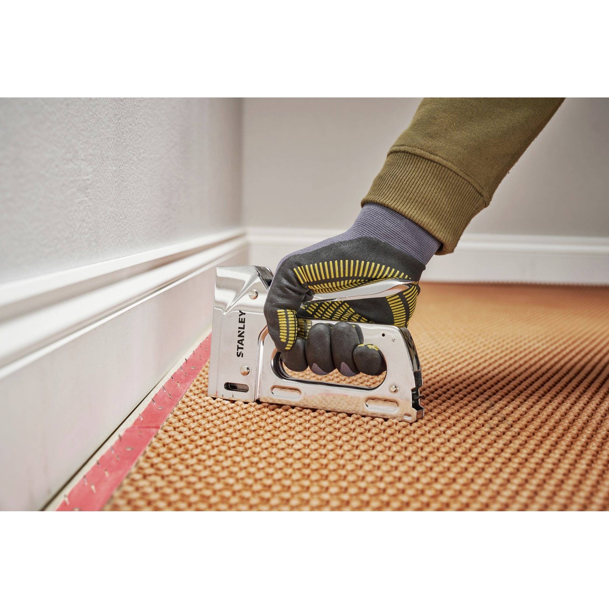 Eine Hand mit Handschuh verwendet einen Tacker, um einen orangefarbenen Teppichboden entlang einer weißen Fußleiste zu befestigen.