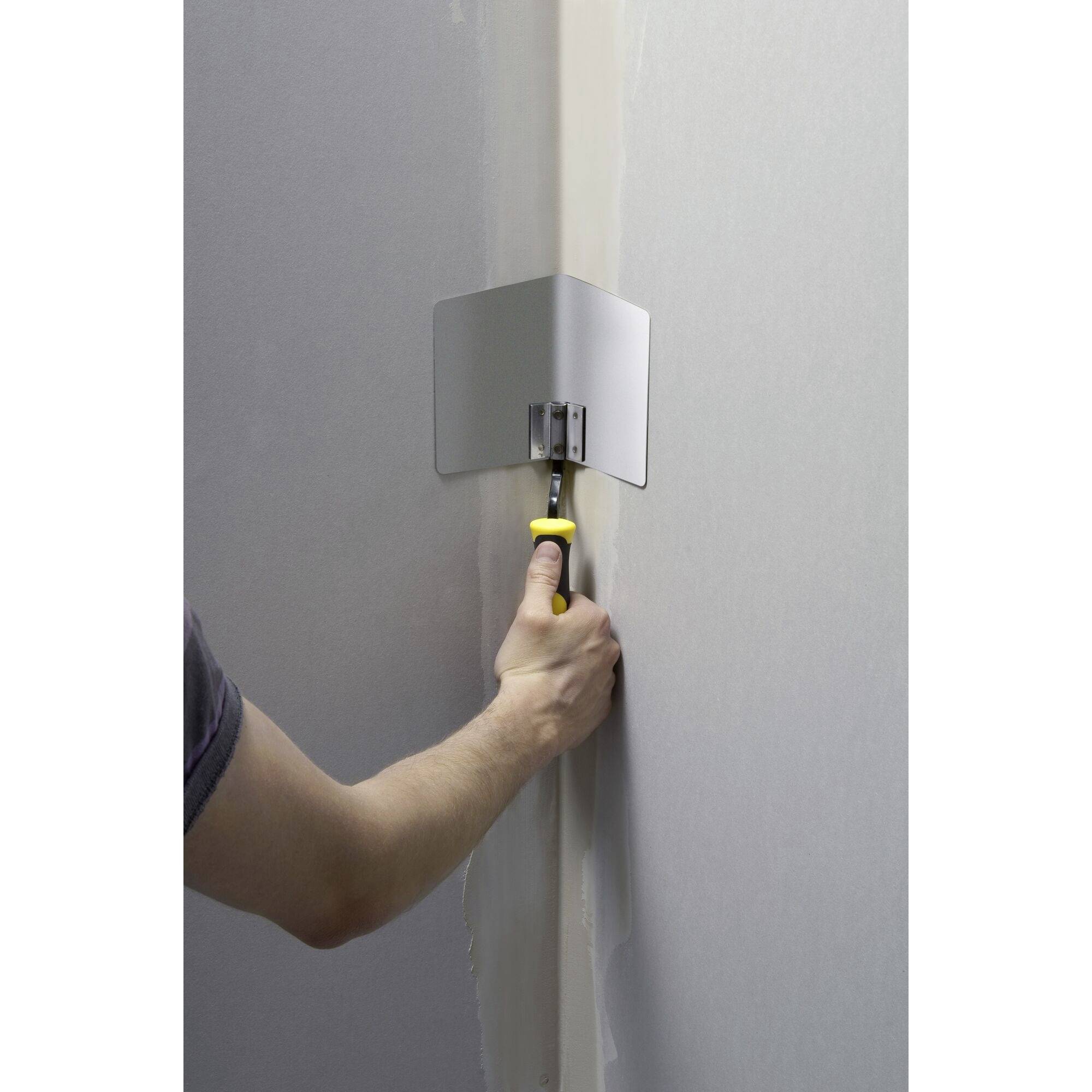 Eine Person hält ein Spachtelwerkzeug an eine Wand, um eine Ecke abzudecken. Das Werkzeug hat einen gelben Griff.