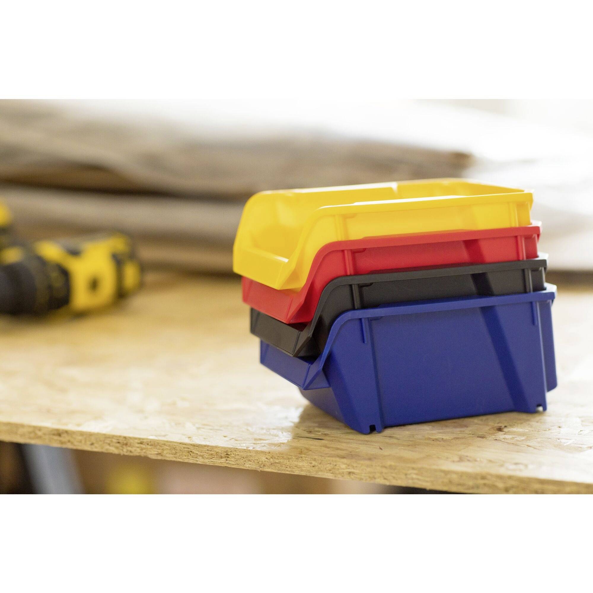 Vier stapelbare Behälter in Gelb, Rot, Schwarz und Blau auf einem Holztisch, im Hintergrund sind unscharfe Werkzeuge sichtbar.