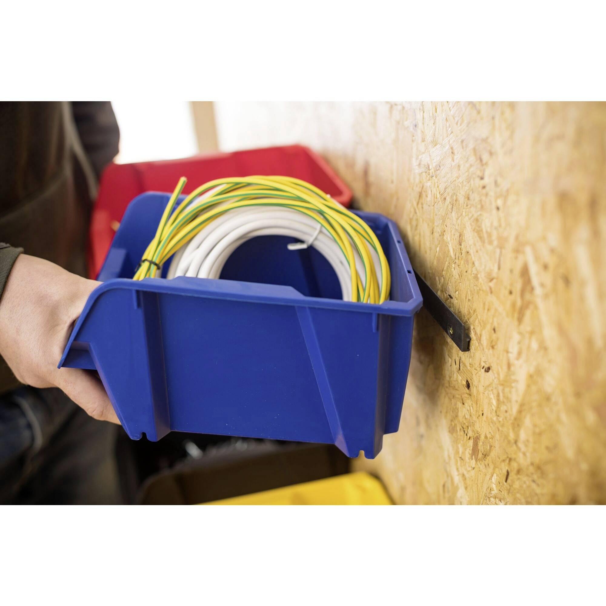 Eine Person hält eine blaue Kiste mit aufgerollten Kabeln vor einer Holzplatte. Im Hintergrund sind zwei weitere Kisten zu sehen.
