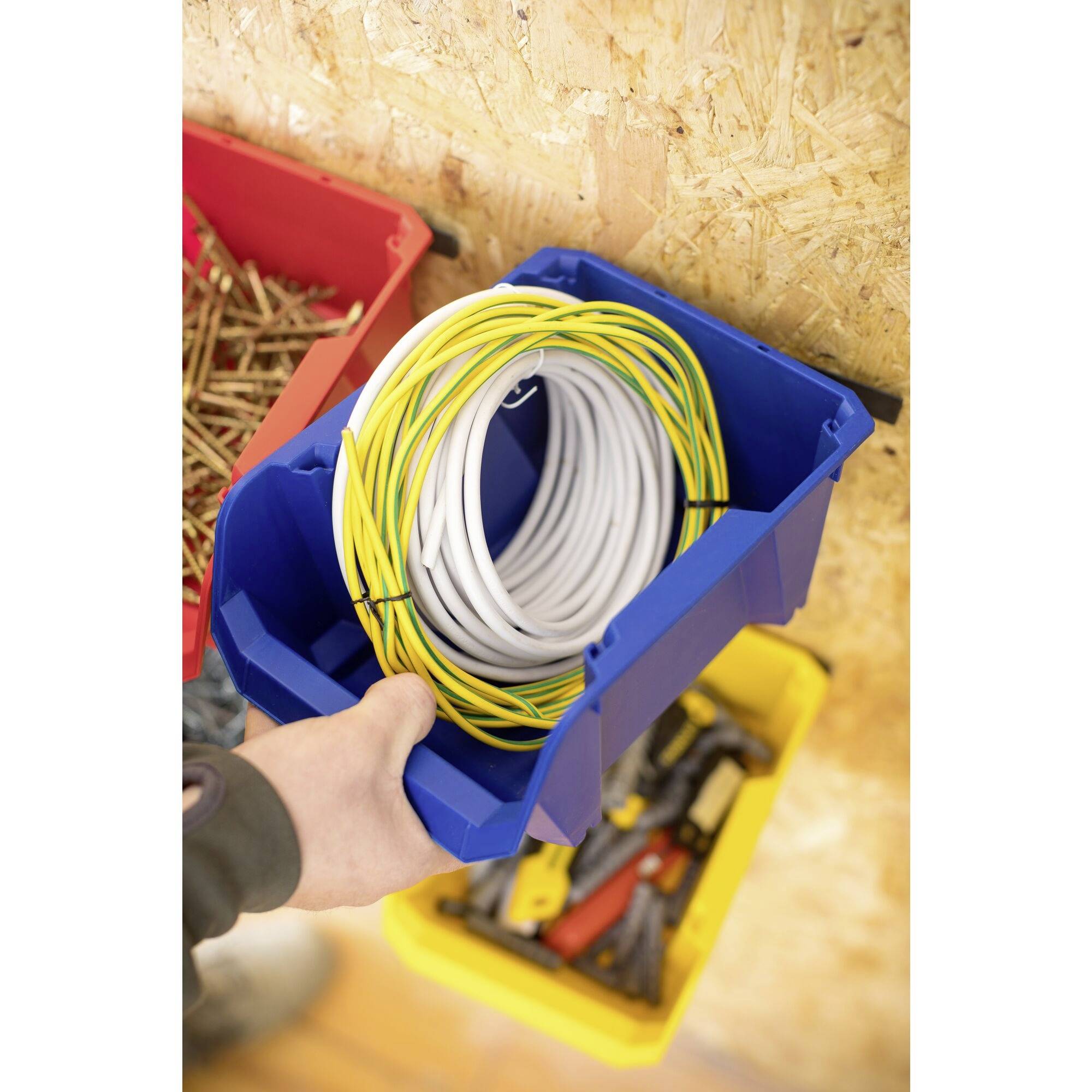 Eine Hand greift nach einer blauen Kiste mit gelben und weißen Kabeln, daneben sind rote und gelbe Kisten mit Schrauben und Werkzeugen.