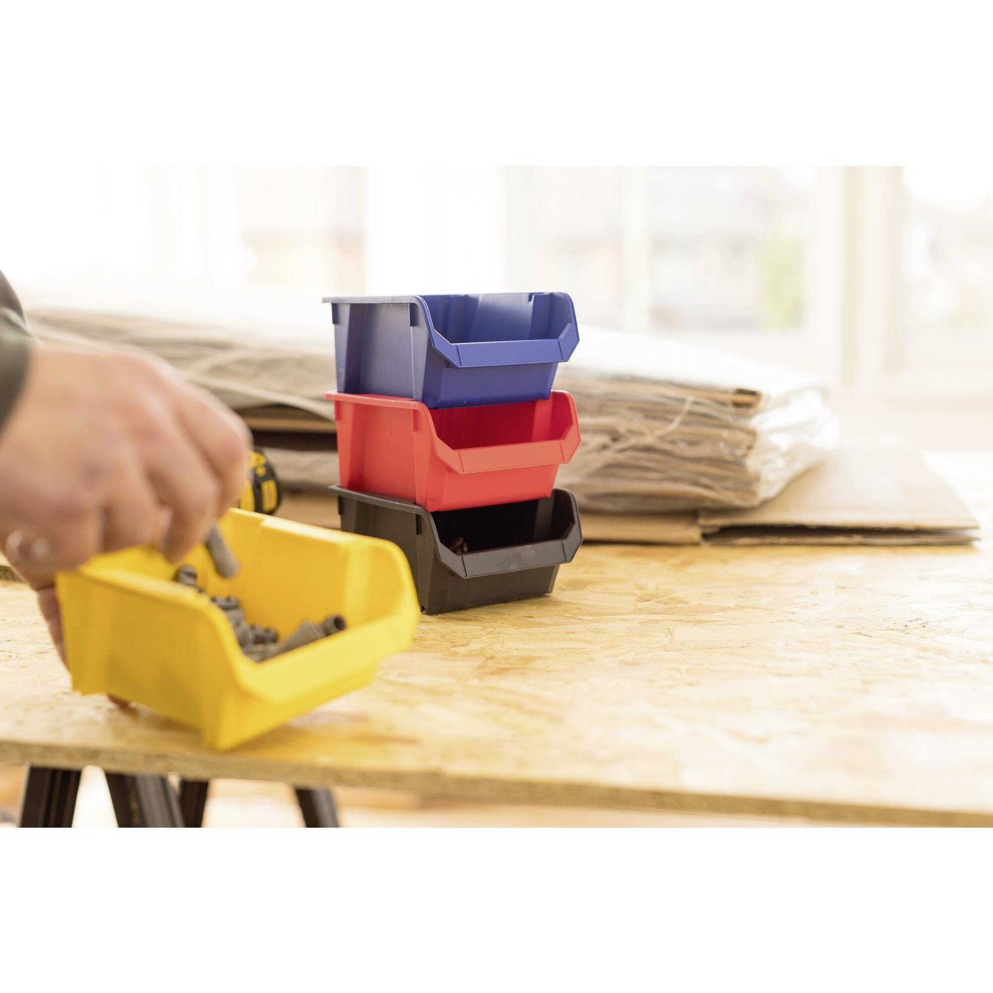 Kleine gestapelte Behälter in Blau, Rot, Schwarz und Gelb stehen auf einem Holztisch. Eine Hand legt Schrauben in den gelben Behälter.