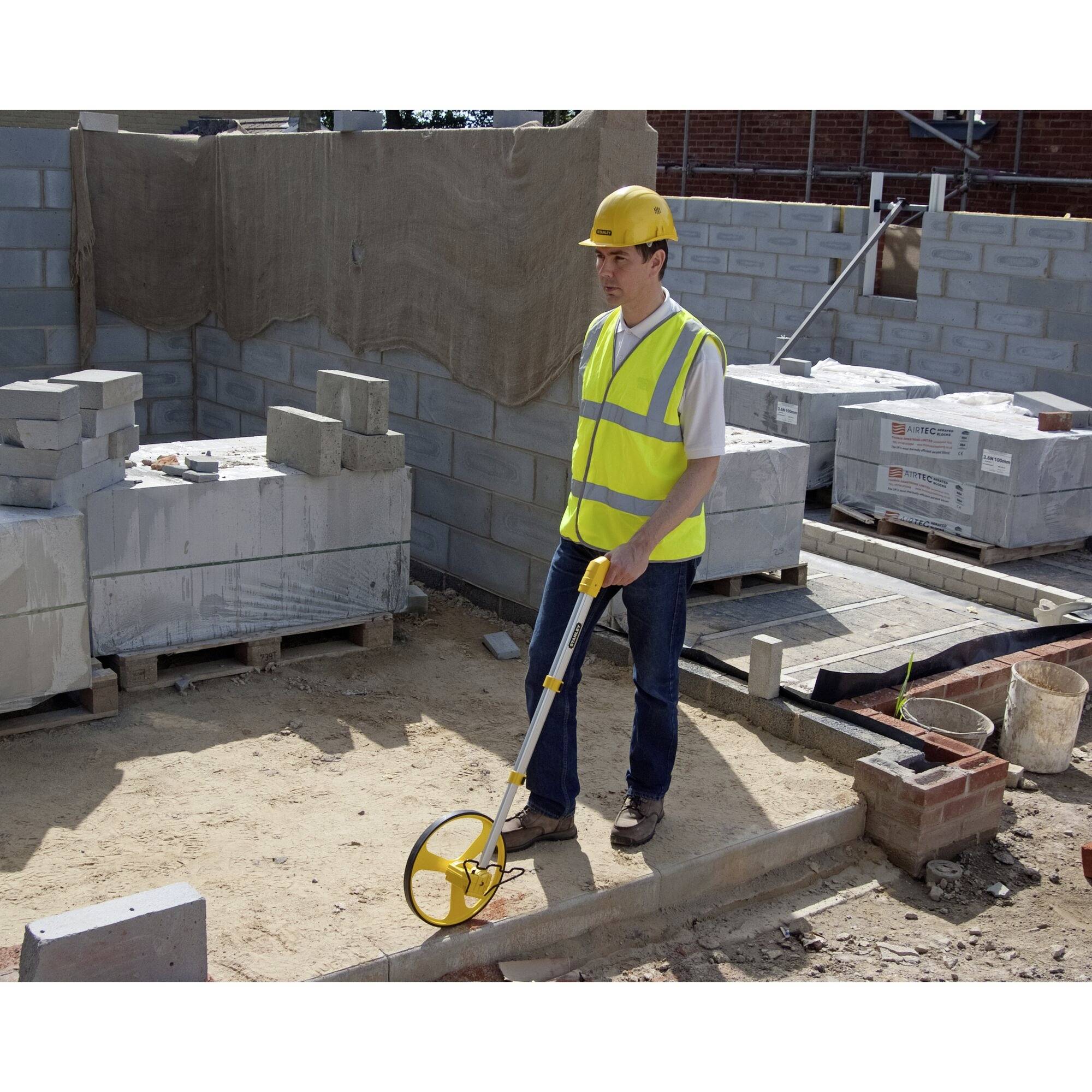 Eine Person auf einer Baustelle misst den Boden mit einem Messrad. Sie trägt einen gelben Schutzhelm und eine Warnweste. Im Hintergrund sind Bauzubehör und unverputzte Wände zu sehen.