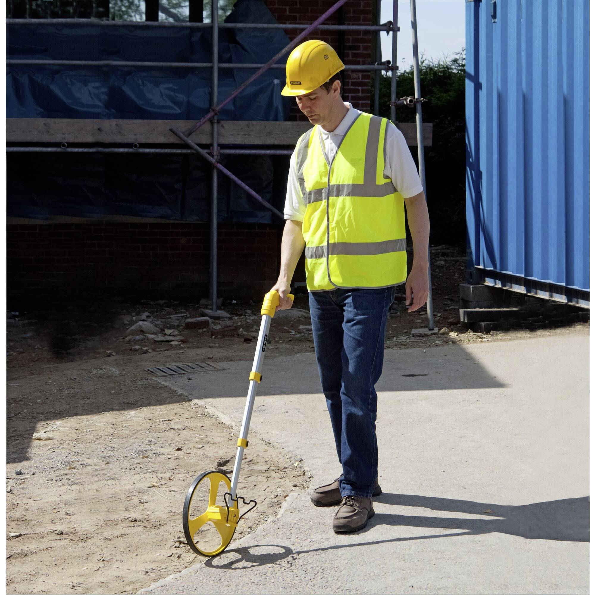 Ein Bauarbeiter misst mit einem Messrad eine Strecke auf einer Baustelle. Er trägt Sicherheitskleidung und einen Schutzhelm.
